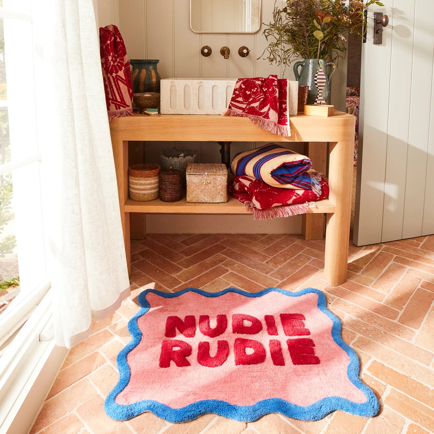 Sage and Clare nudie rudie bath mat with wavy edge in blue, on peachy pink back ground with red "Nudie Rudie" text