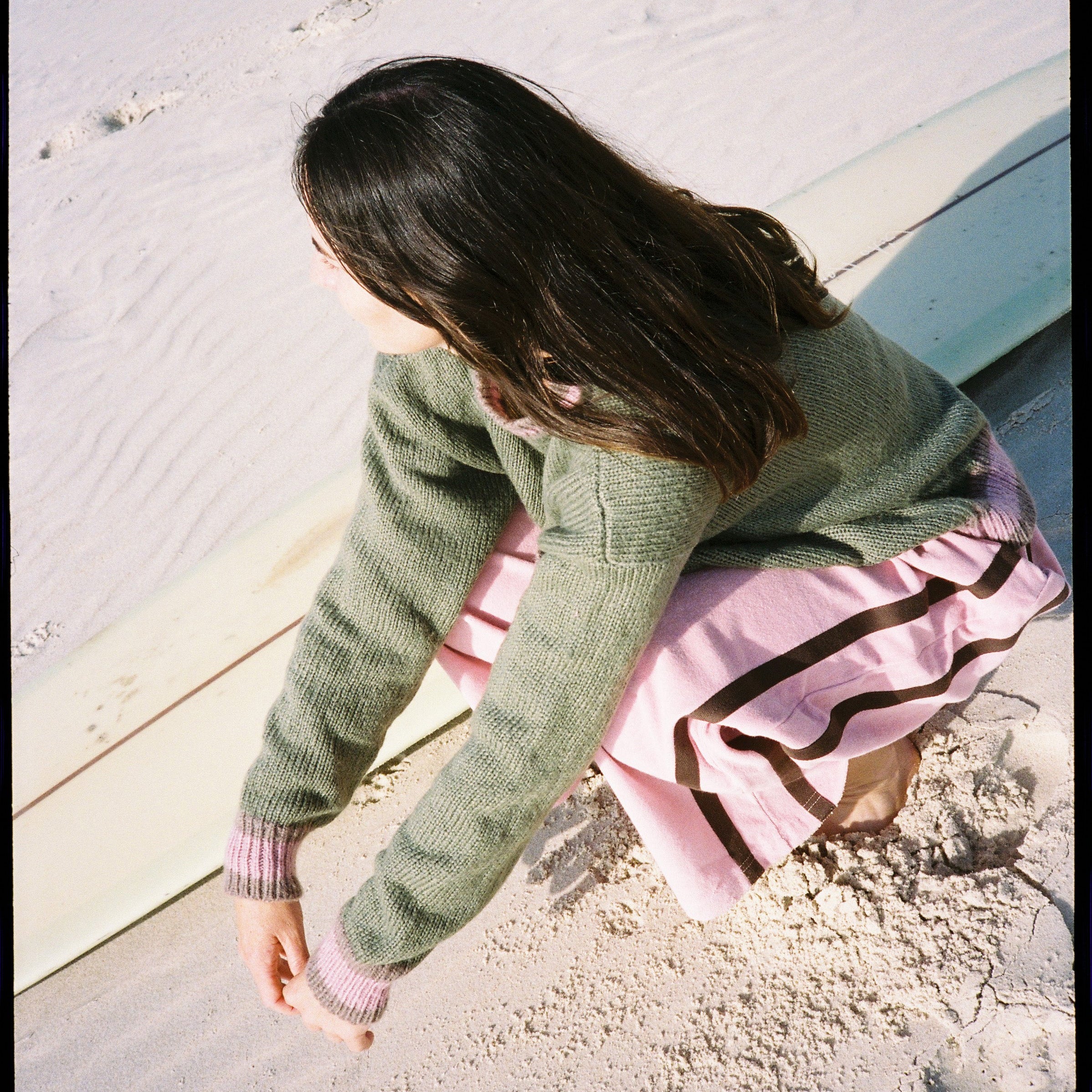 Overhead angle of model in blush pink Après Surf Panel Pants sitting on beach with surfboard