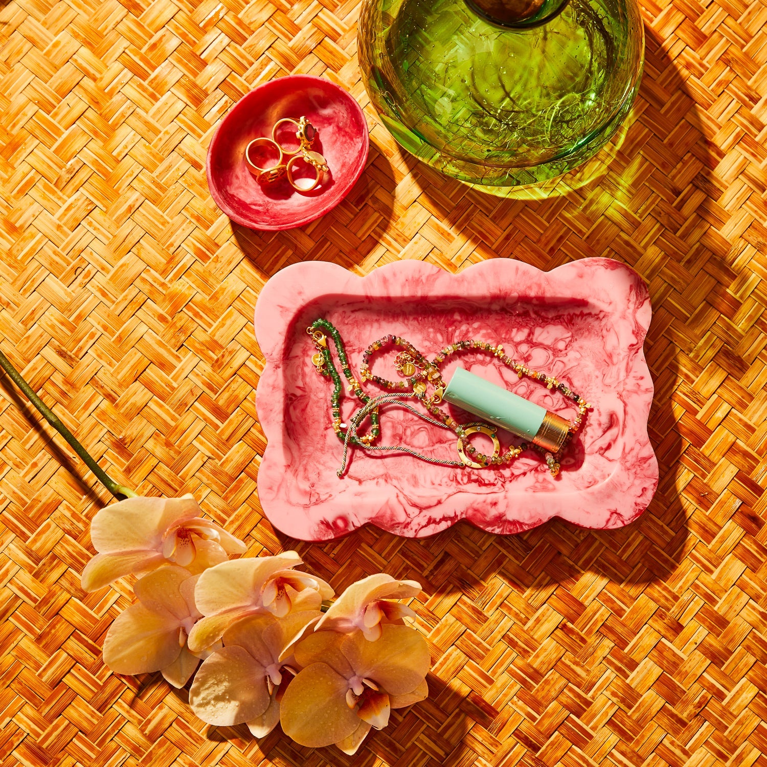 Crimson marbled resin small tray holding jewelry and lipstick, styled on a woven rattan surface with orchids