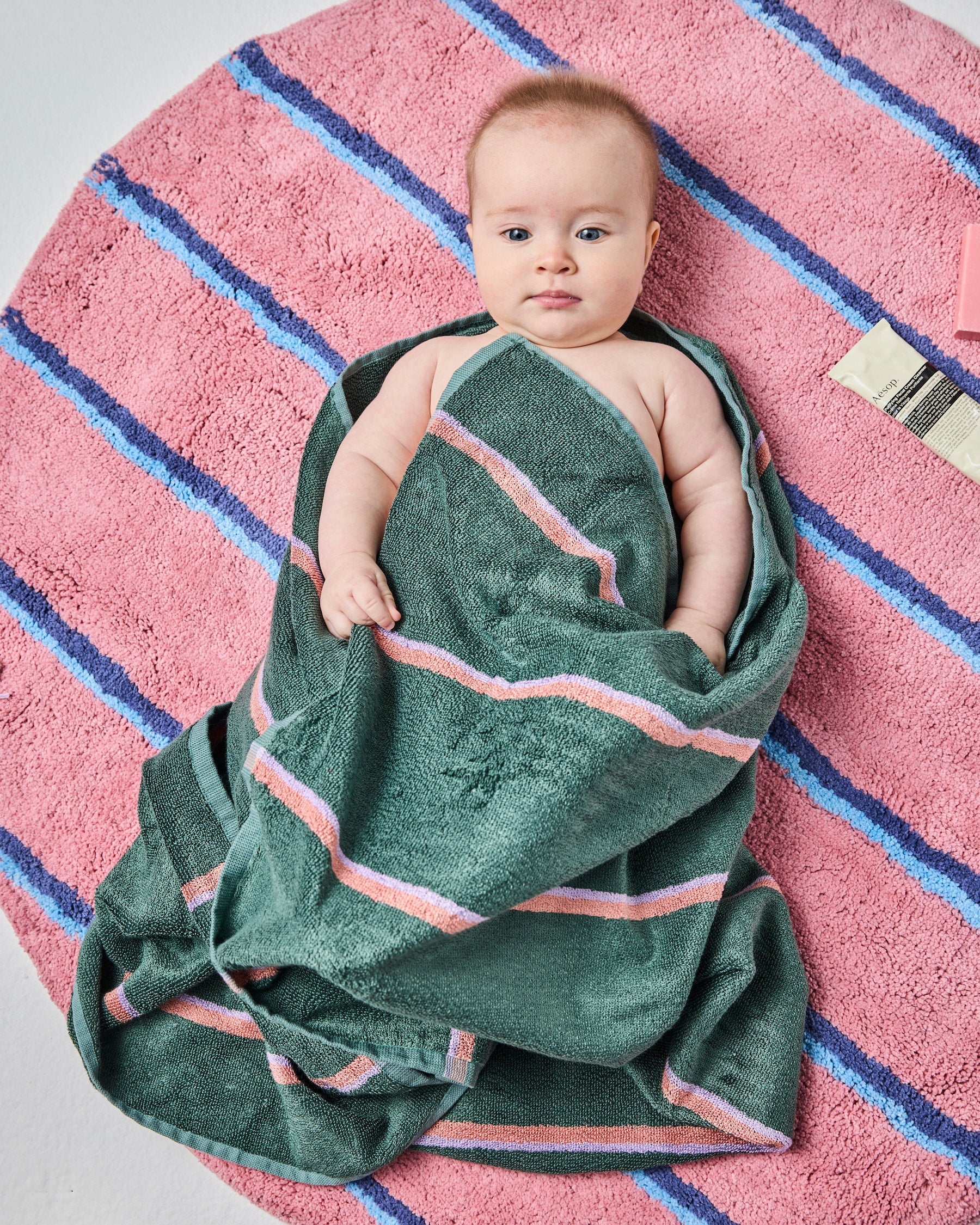 Baby lying on Blushing Stripe Round Bath Mat by Kip & Co wrapped in green striped towel showing softness