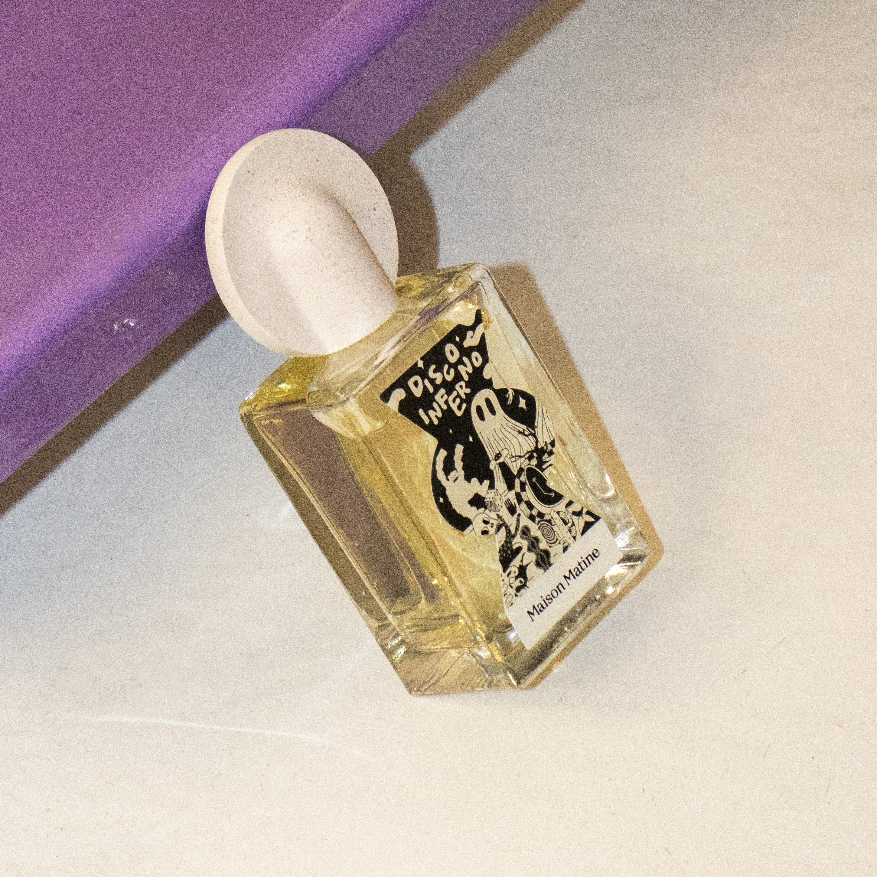 Angled view of Disco Inferno fragrance bottle with round pink cap on purple and cream geometric surface