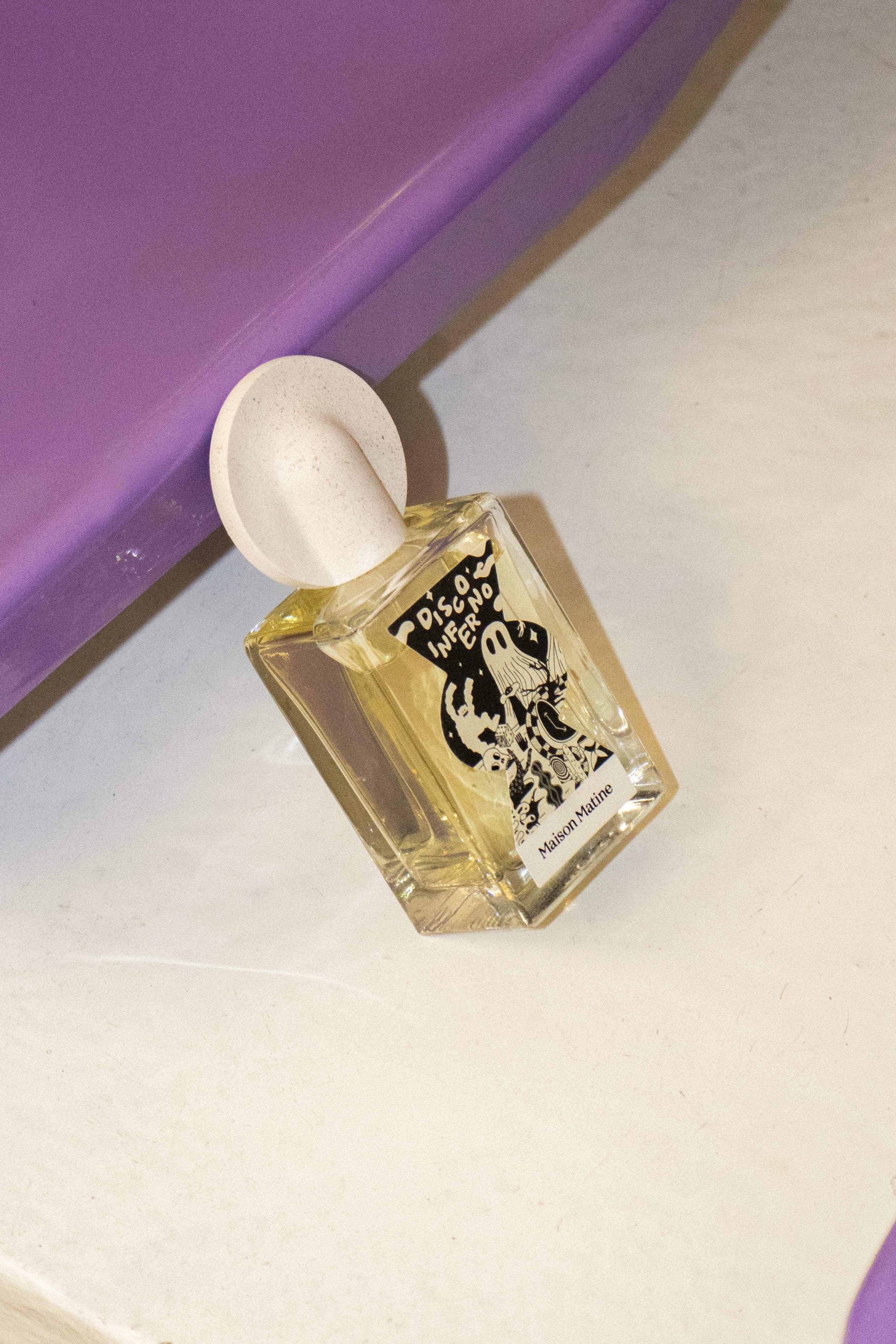 Angled view of Disco Inferno fragrance bottle with round pink cap on purple and cream geometric surface