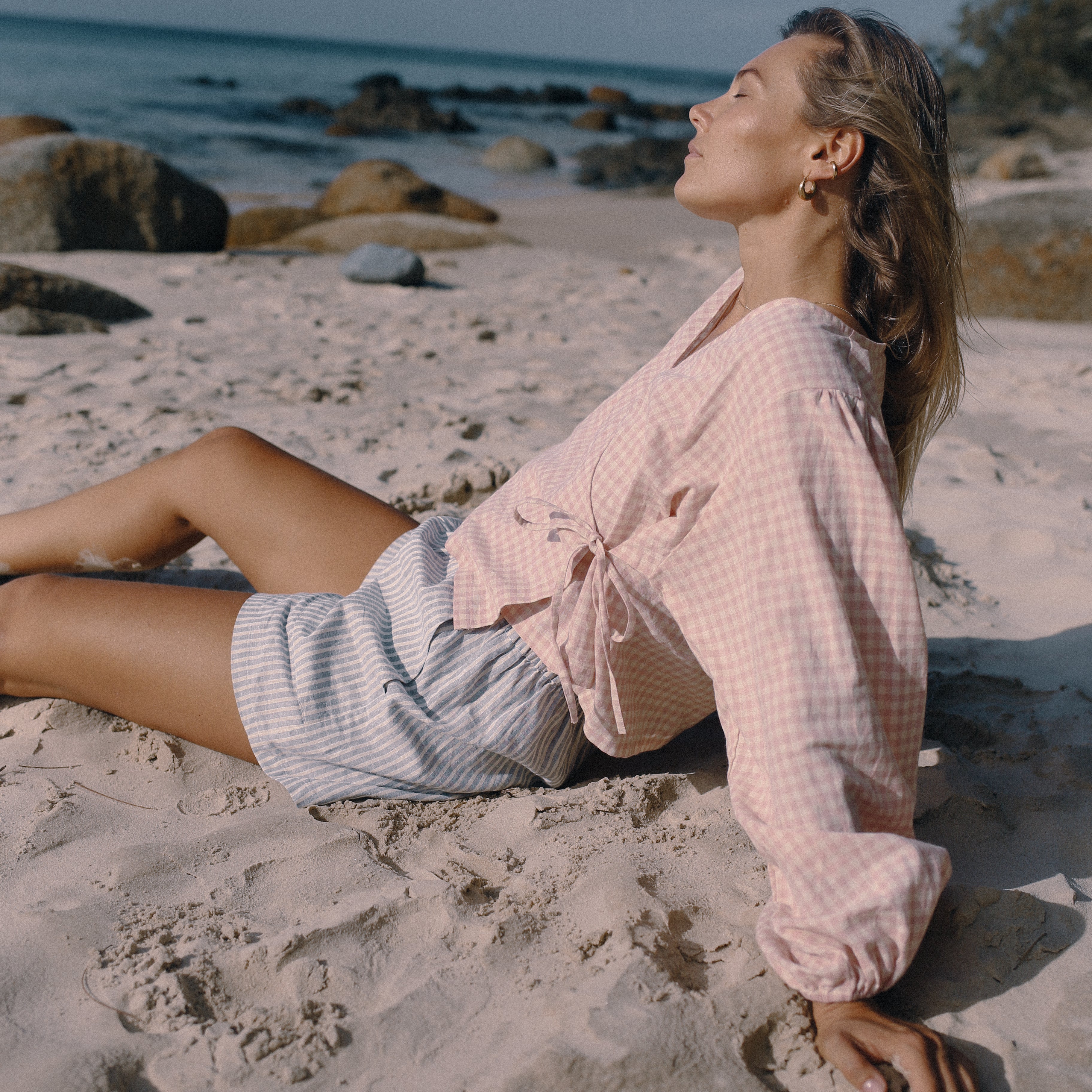 Woman in a pink shirt and white shorts sitting on a sandy beach with ocean and rocks in the background.