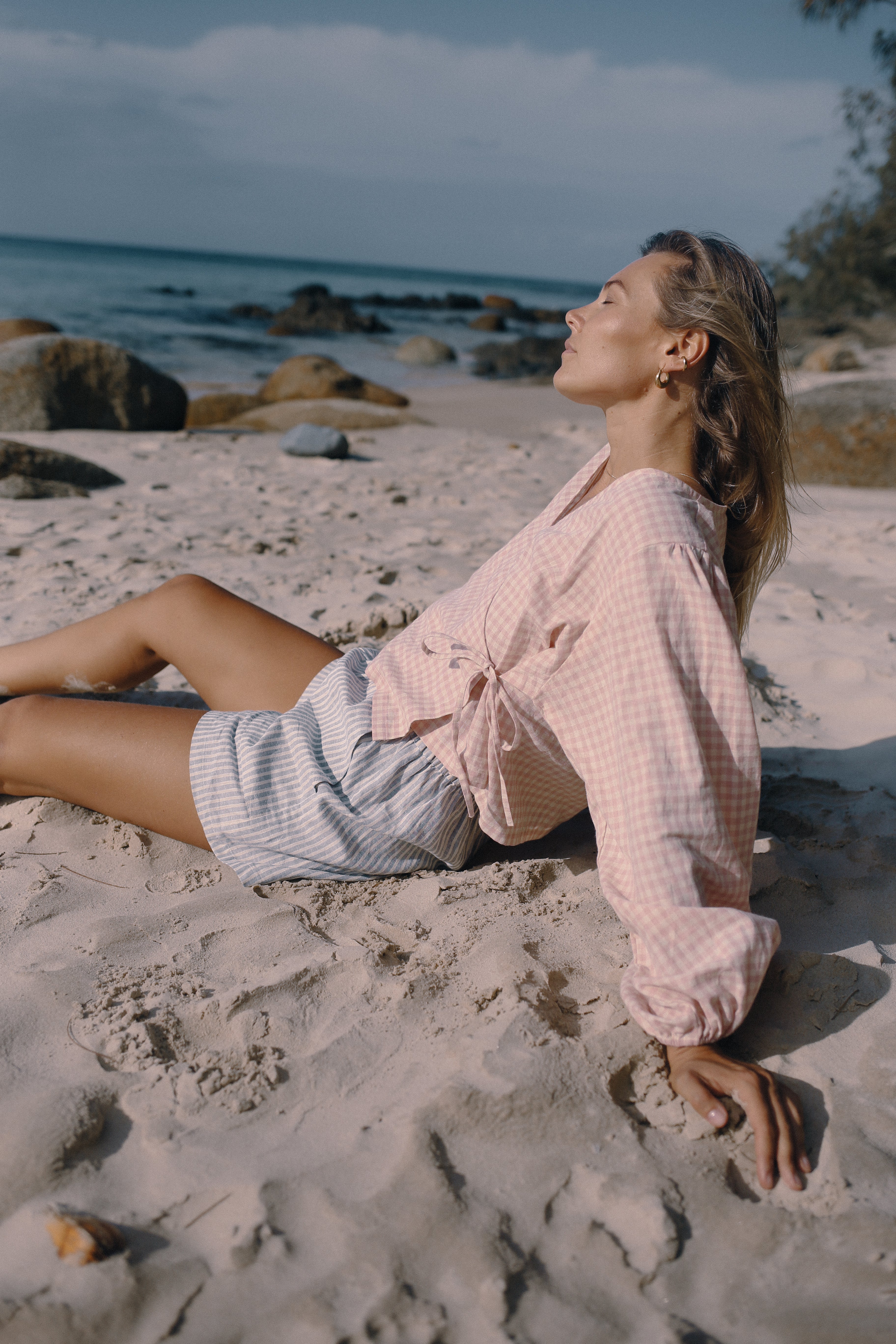 Woman in a pink shirt and white shorts sitting on a sandy beach with ocean and rocks in the background.