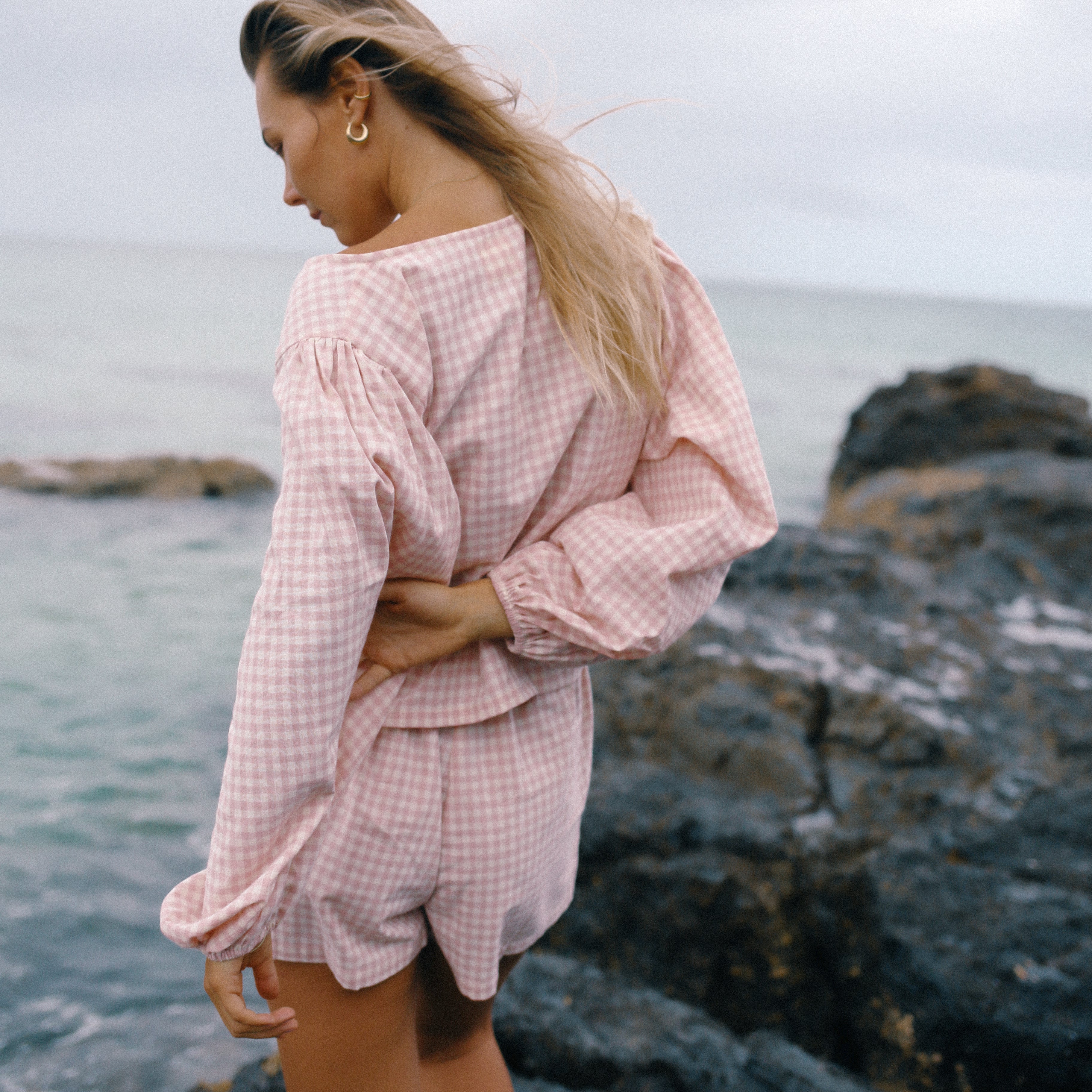 Woman in a pink checkered outfit standing on rocky terrain by the sea wearing the Ella Wrap Top pink gingham by The Bare Road