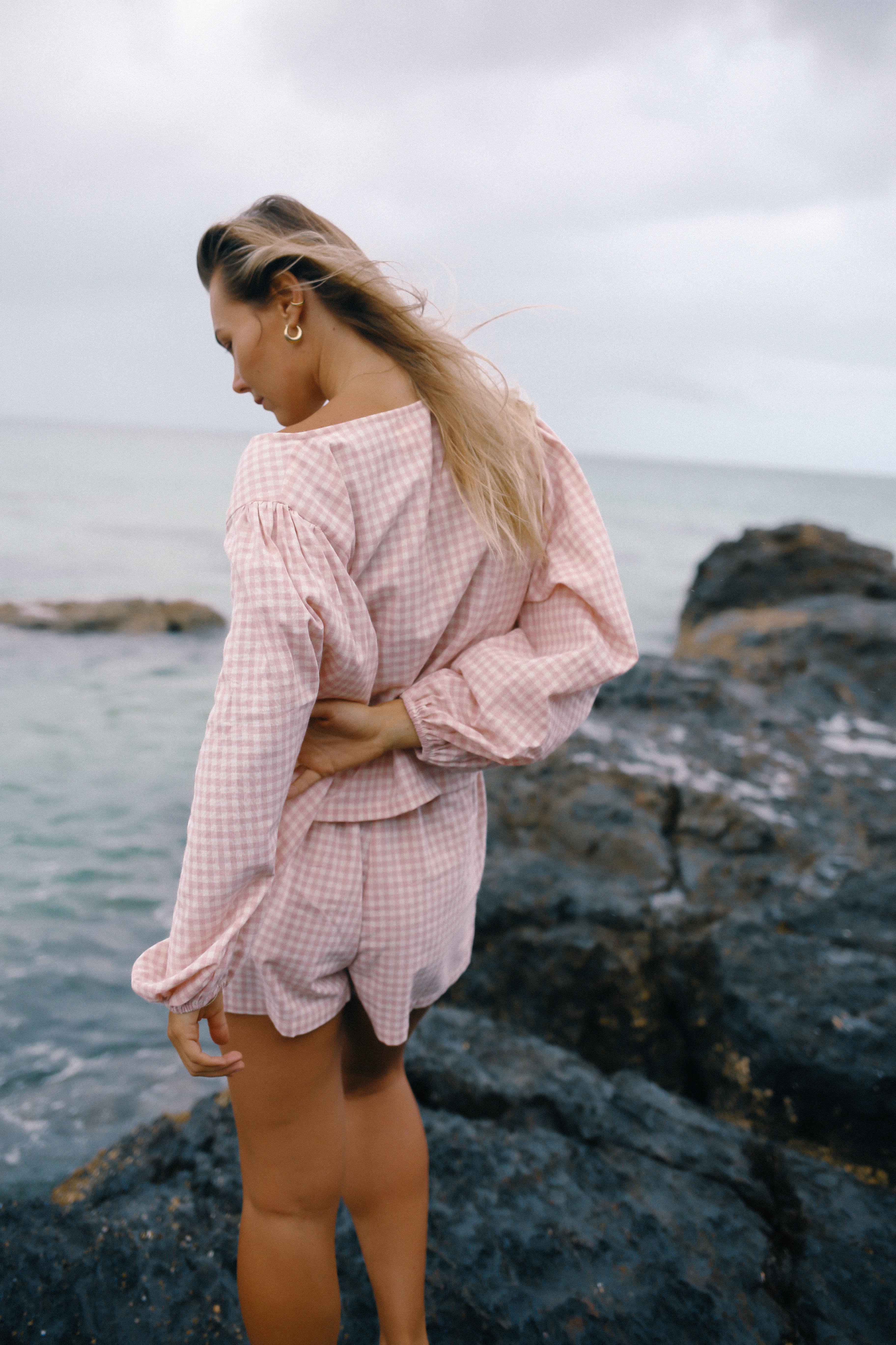 Woman in a pink checkered outfit standing on rocky terrain by the sea wearing the Ella Wrap Top pink gingham by The Bare Road