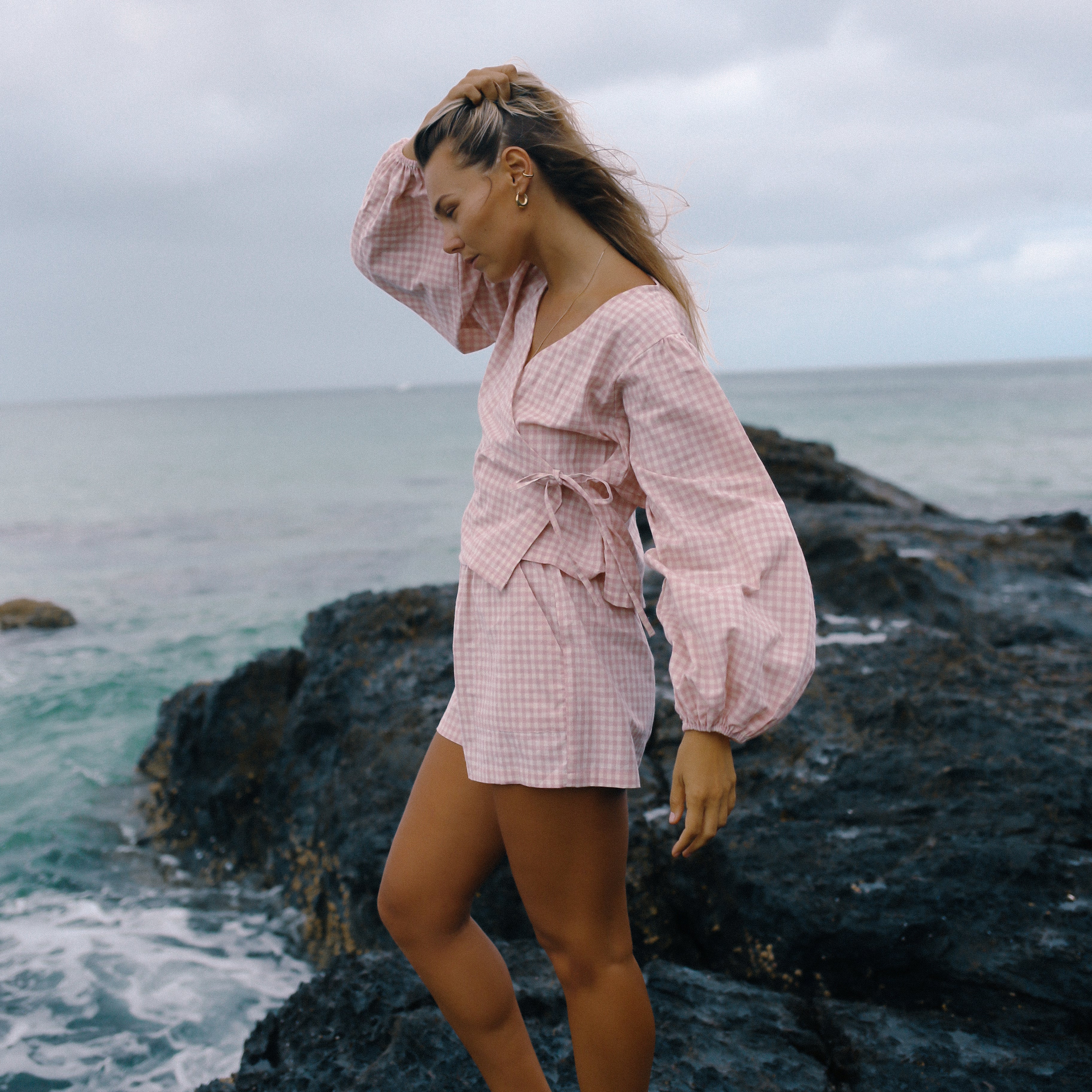 Woman in a pink outfit standing on a rocky outcrop by the ocean under a cloudy sky. Ella Wrap Top pink gingham by The Bare Road