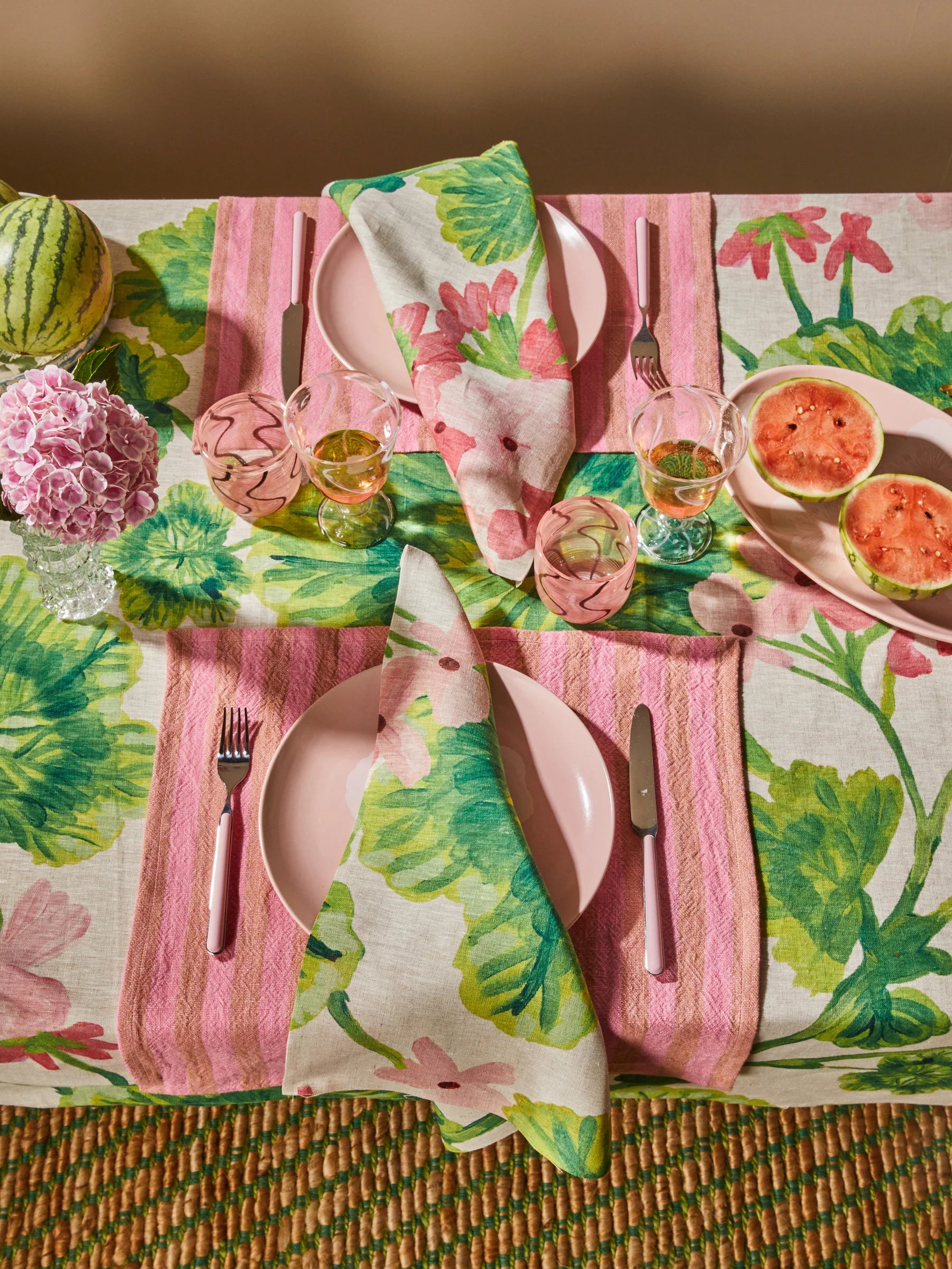 Colorful Bonnie & Neil table setting with floral tablecloth, pink plates, and watermelon on a woven surface.