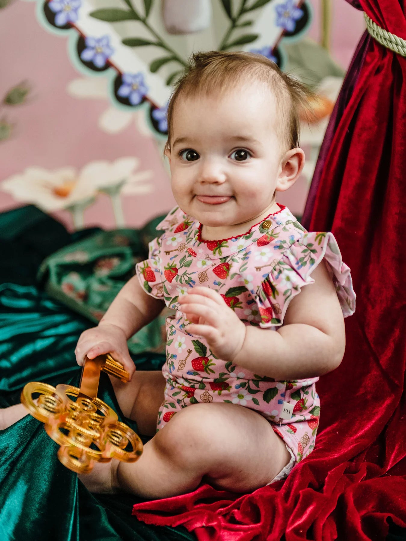 Baby in a floral outfit holding a toy, sitting on a red and green fabric with a floral-patterned background.