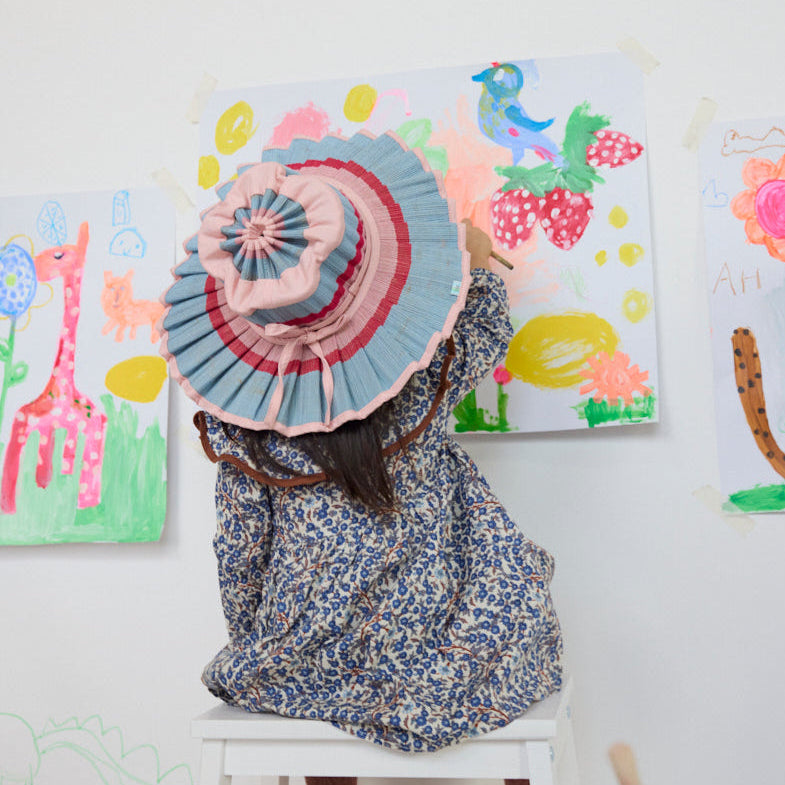 Side lifestyle shot of child in Luxe Capri Hat Ice Cream style sitting against colorful artwork wall
