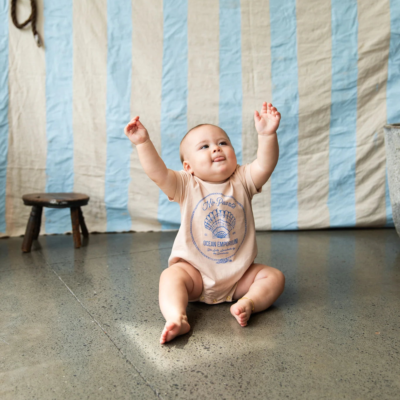 Baby wearing cream colours onesie with blue shell graphic "Miss Pearls Ocean Emporium" by Pearson & Pearson