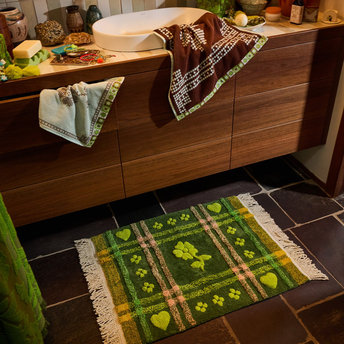 Green tufted bath mat with plaid and floral design styled on a dark tile bathroom floor beside a wooden vanity