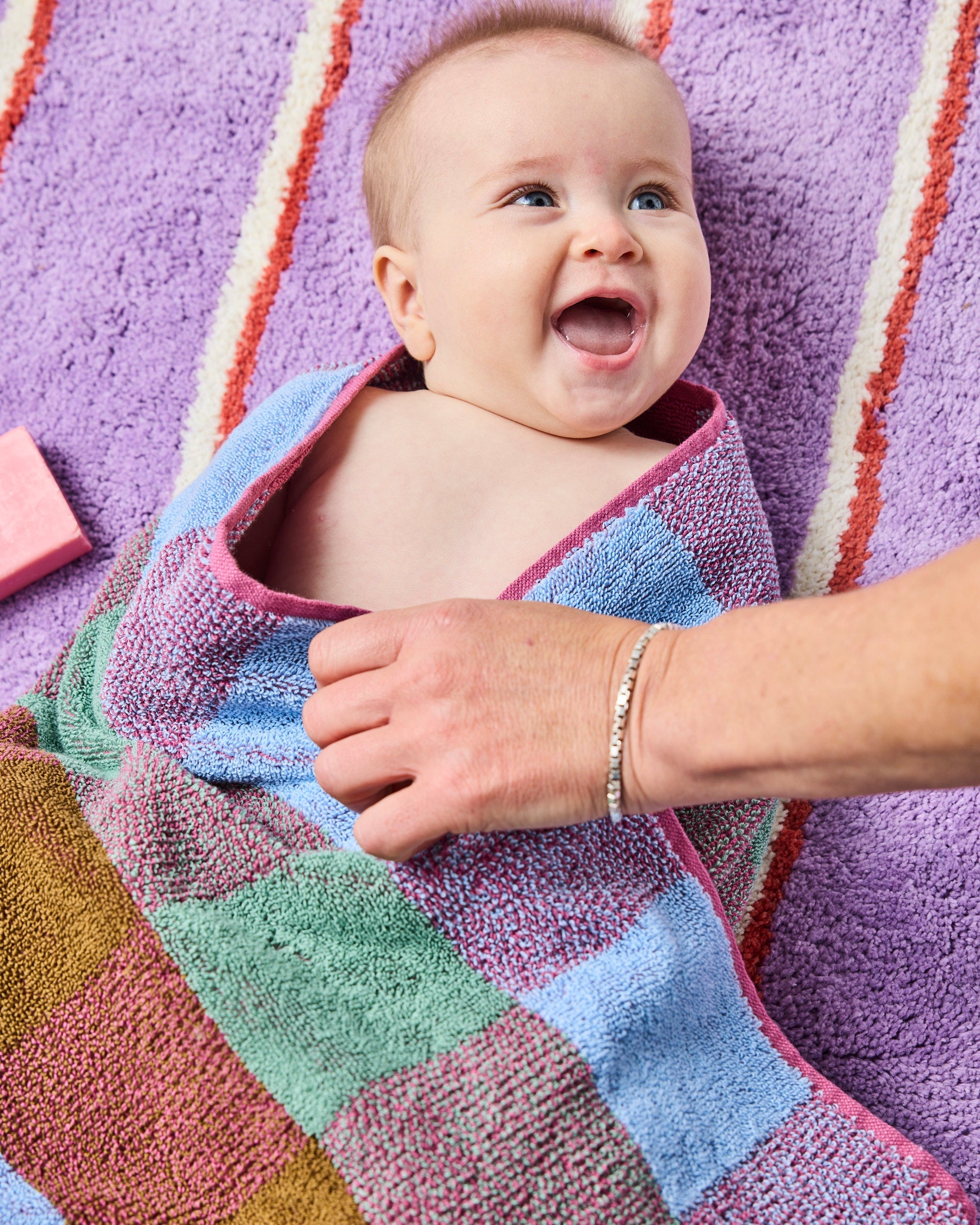 Baby wrapped in colorful striped towel on Lavender Field Stripe Bath Mat showing soft texture and vibrant colors