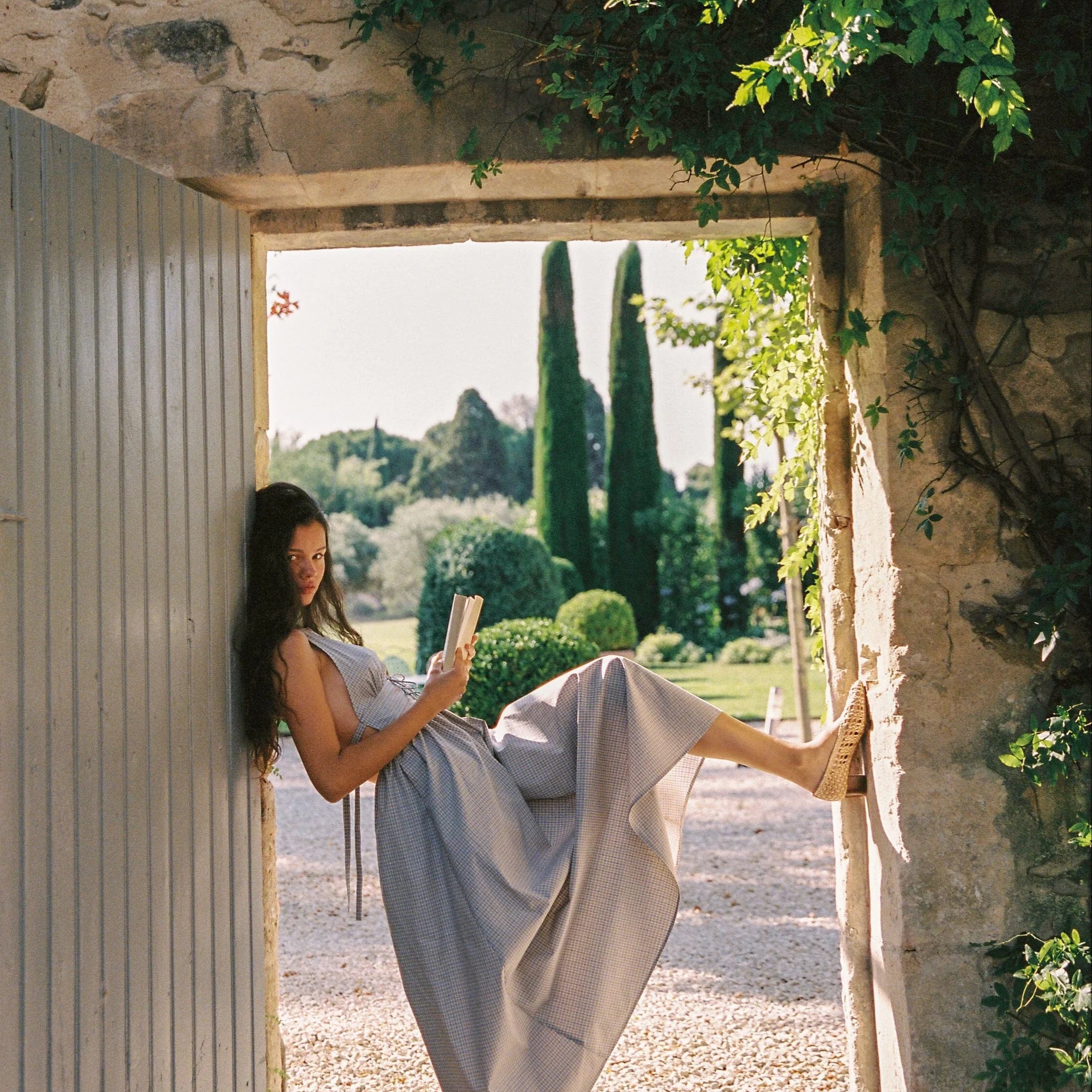 Woman reading a book in a scenic outdoor setting with stone walls and greenery.