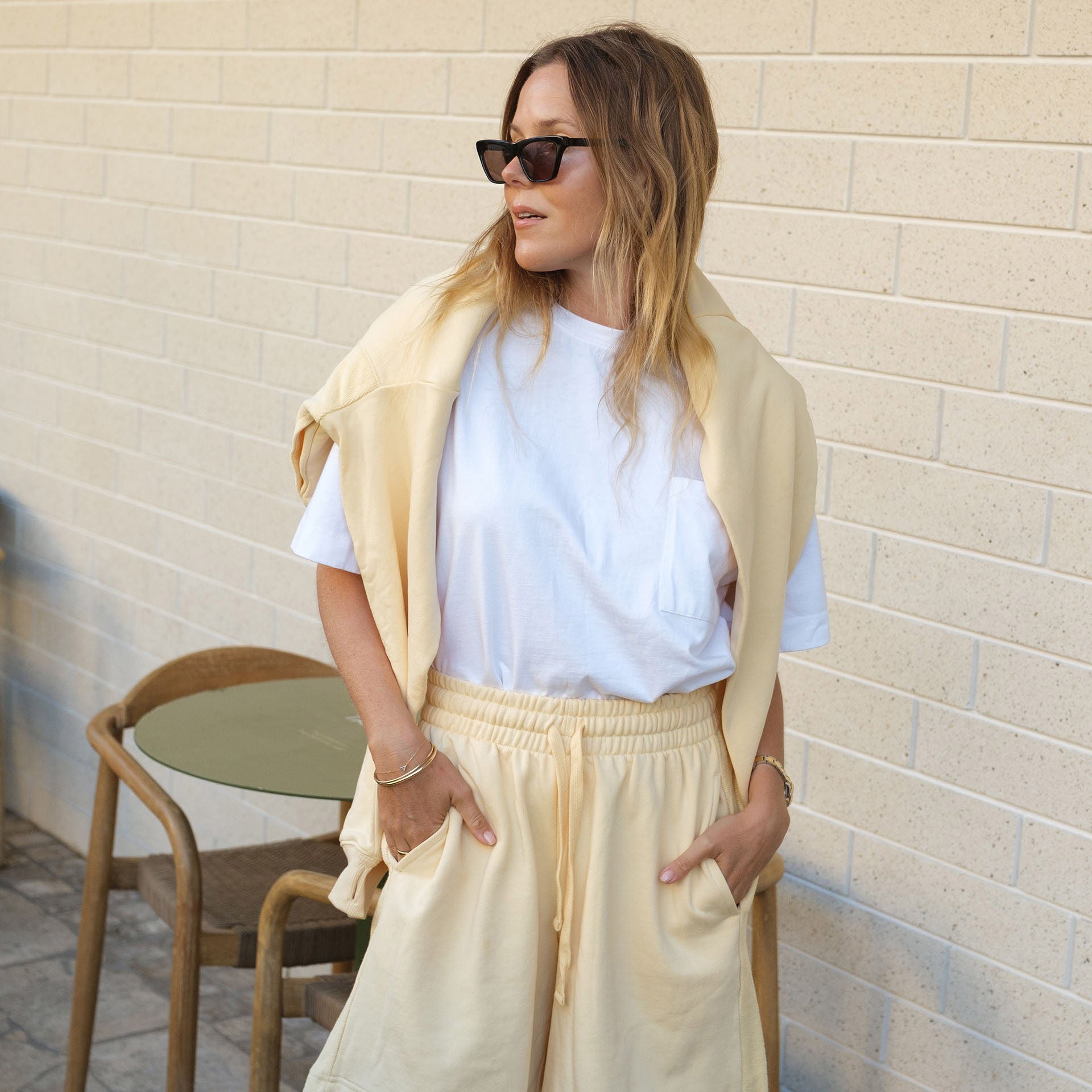 Woman in a light yellow Ceres Life outfit standing against a beige wall with a chair in the background.