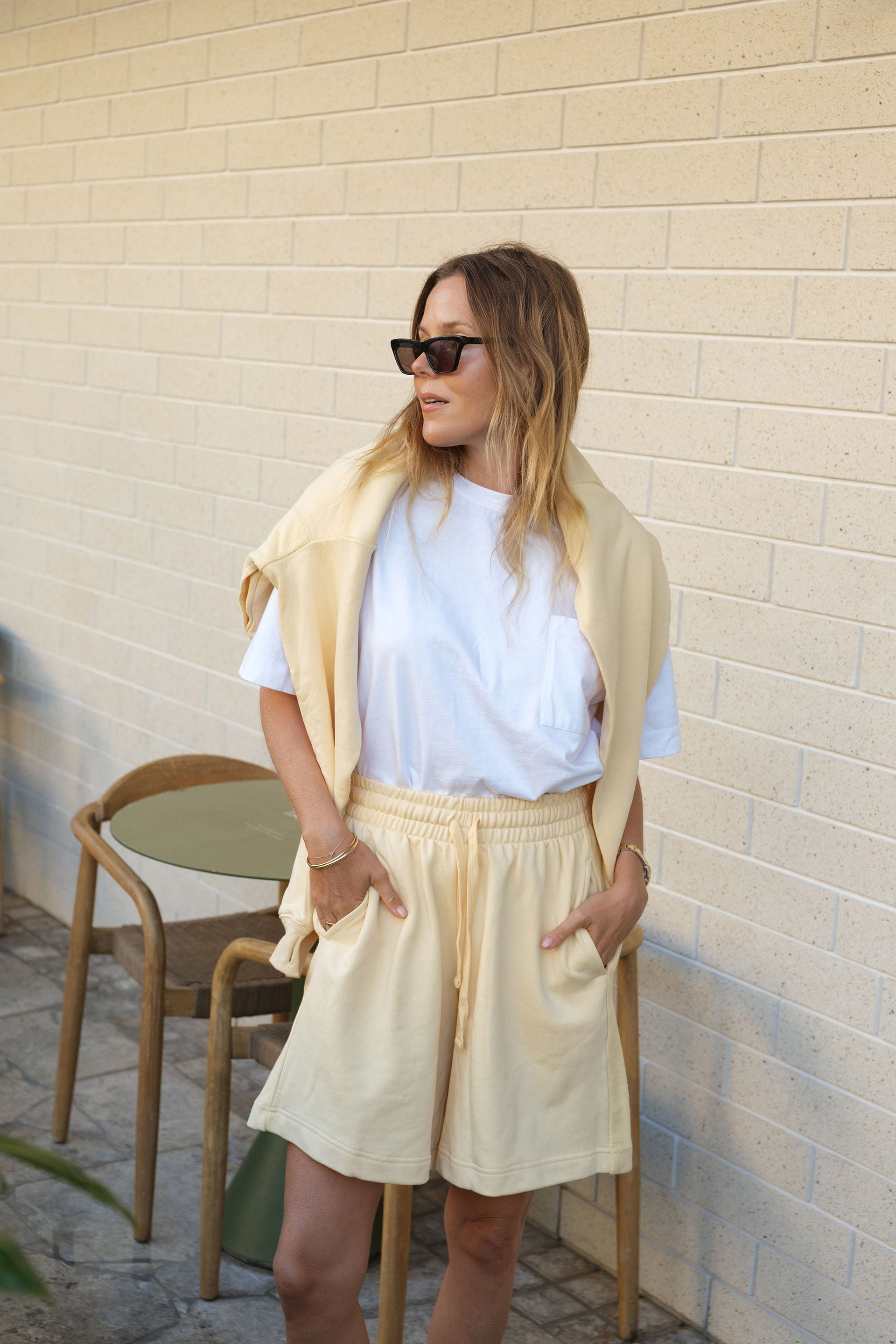 Woman in a light yellow Ceres Life outfit standing against a beige wall with a chair in the background.