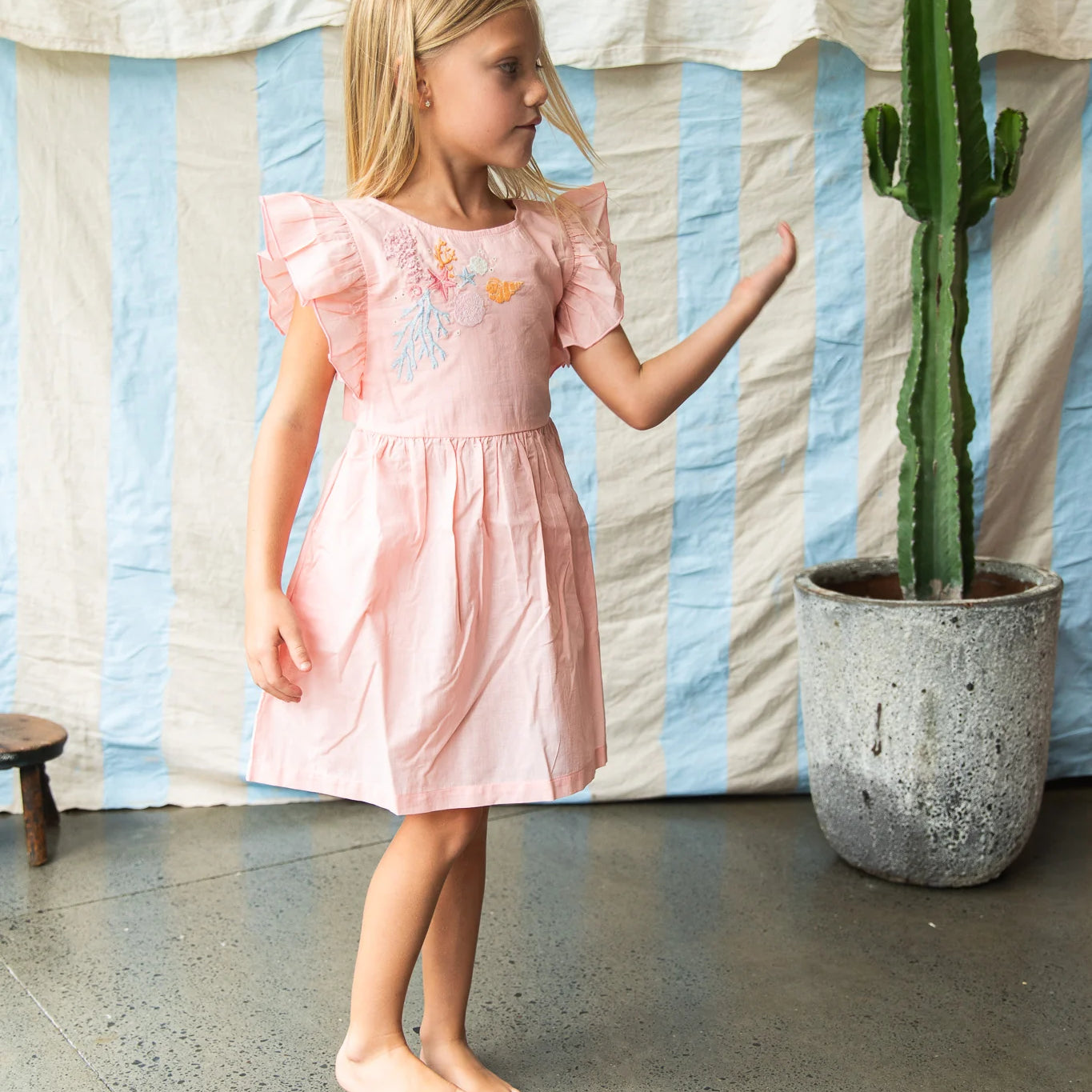 Young girl in a pink dress standing next to a potted cactus indoors.