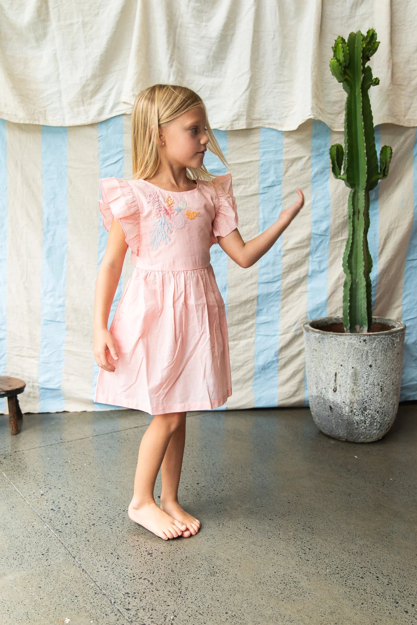 Young girl in a pink dress standing next to a potted cactus indoors.