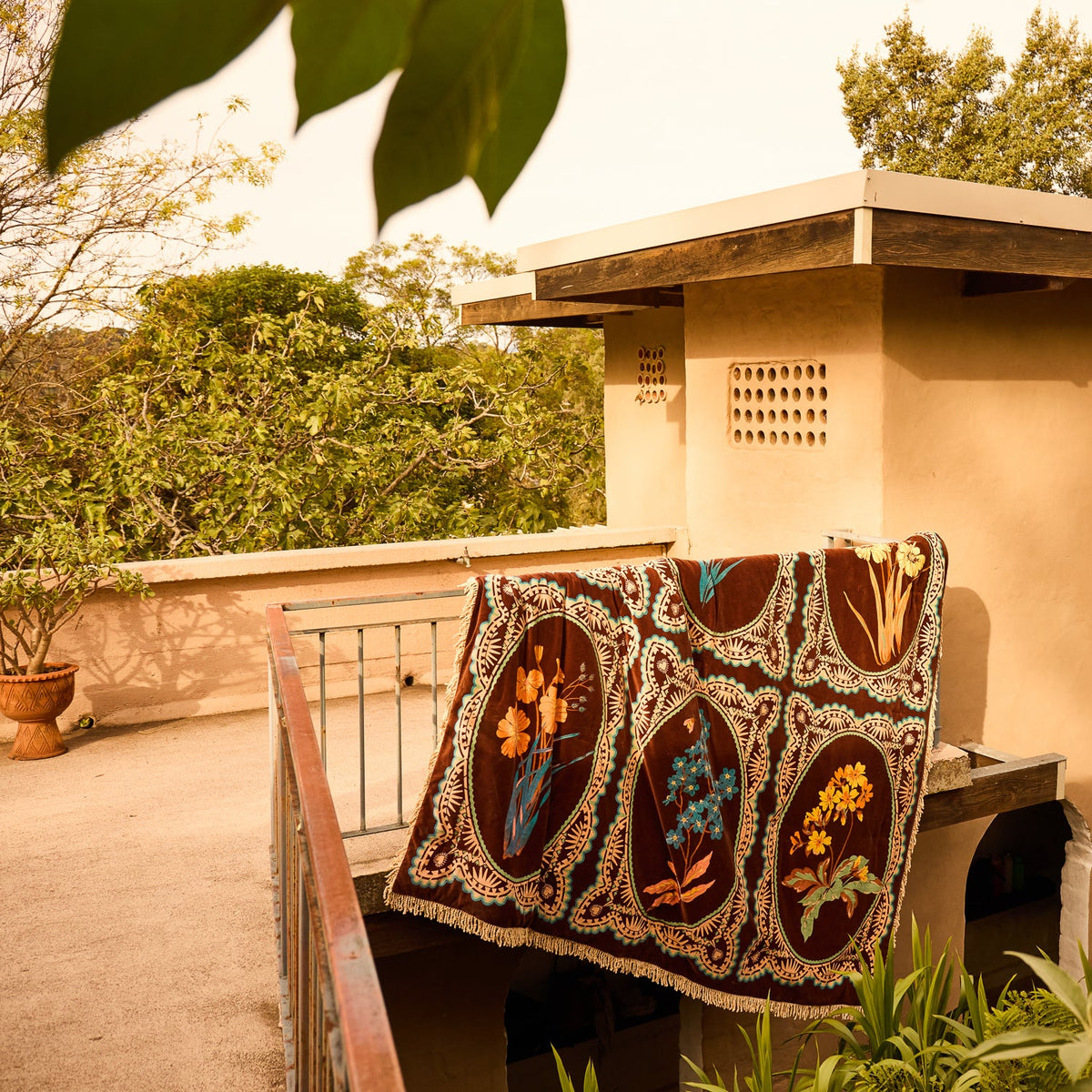 Brown velvet bedcover with six botanical floral medallion panels and cream fringe trim draped over a balcony railing outdoors
