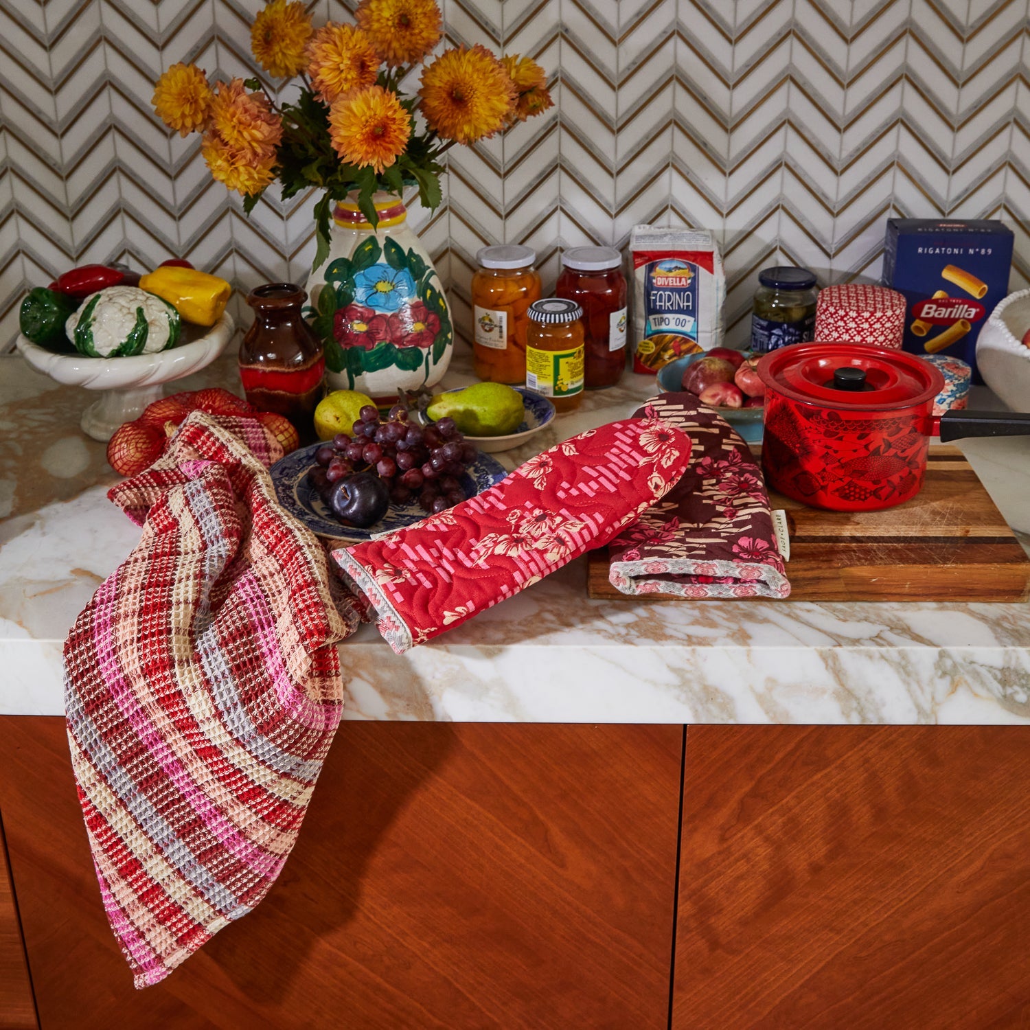 Kitchen styling scene with Marta Oven Mitts on wooden cutting board alongside red cookware and fresh flowers