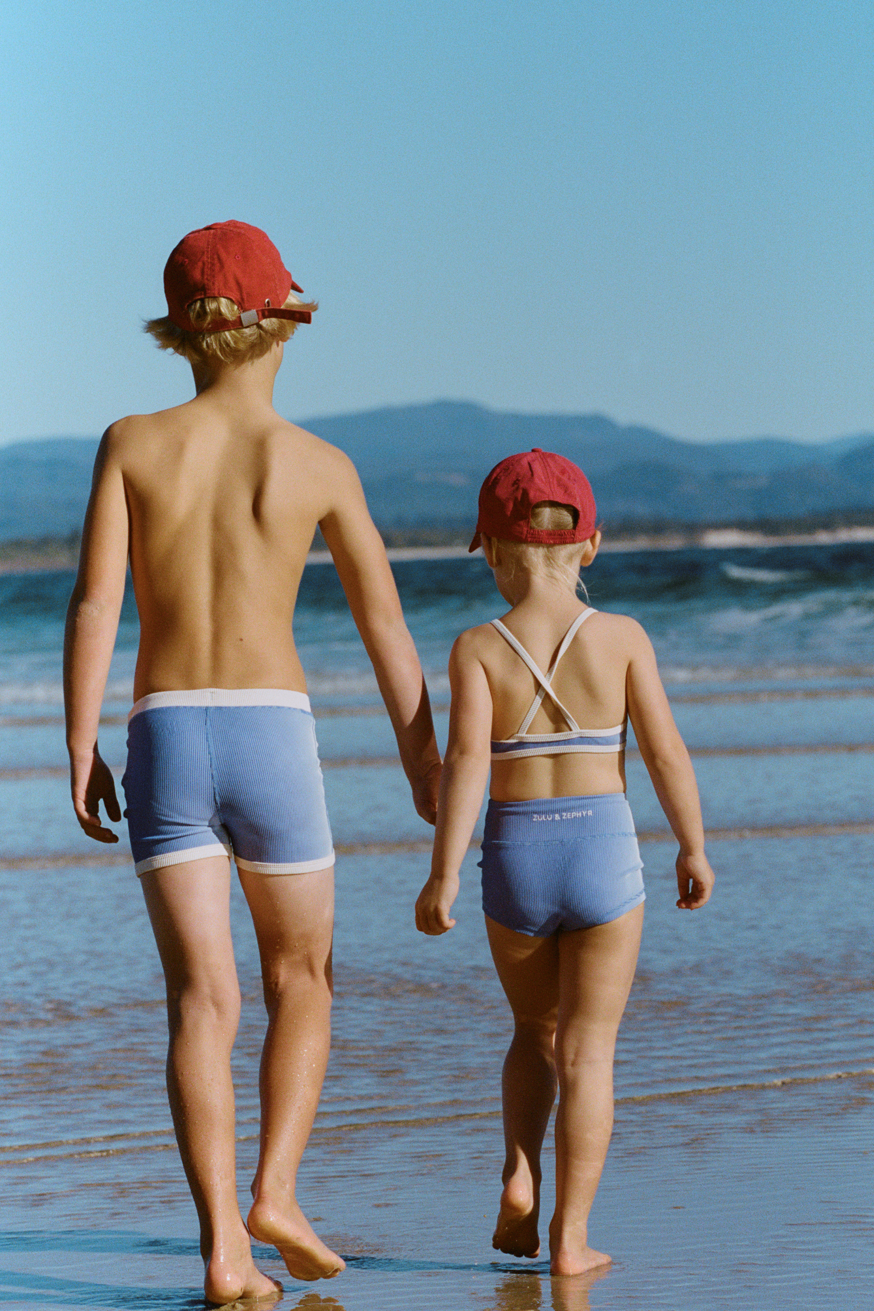 Two children walking on the beach wearing blue mini stripe surf shorts and matching swimwear, rear view