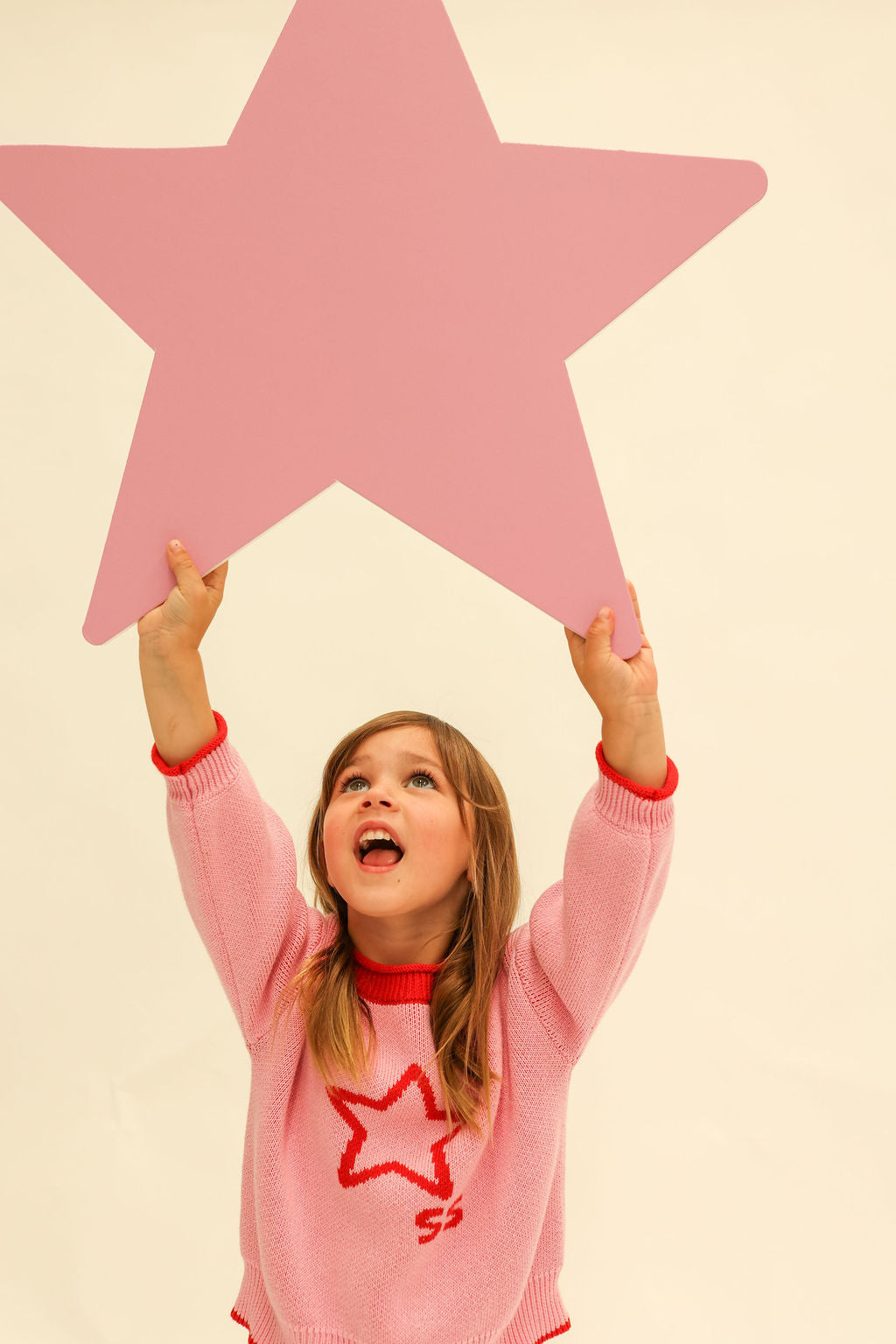 Child wearing pink Neve Knit sweater with red star motif reaching up toward large pink star prop