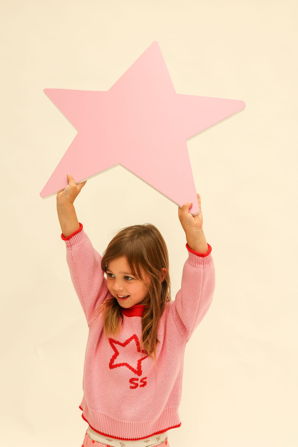 Child in pink Neve Knit sweater holding large pink star prop overhead with arms raised