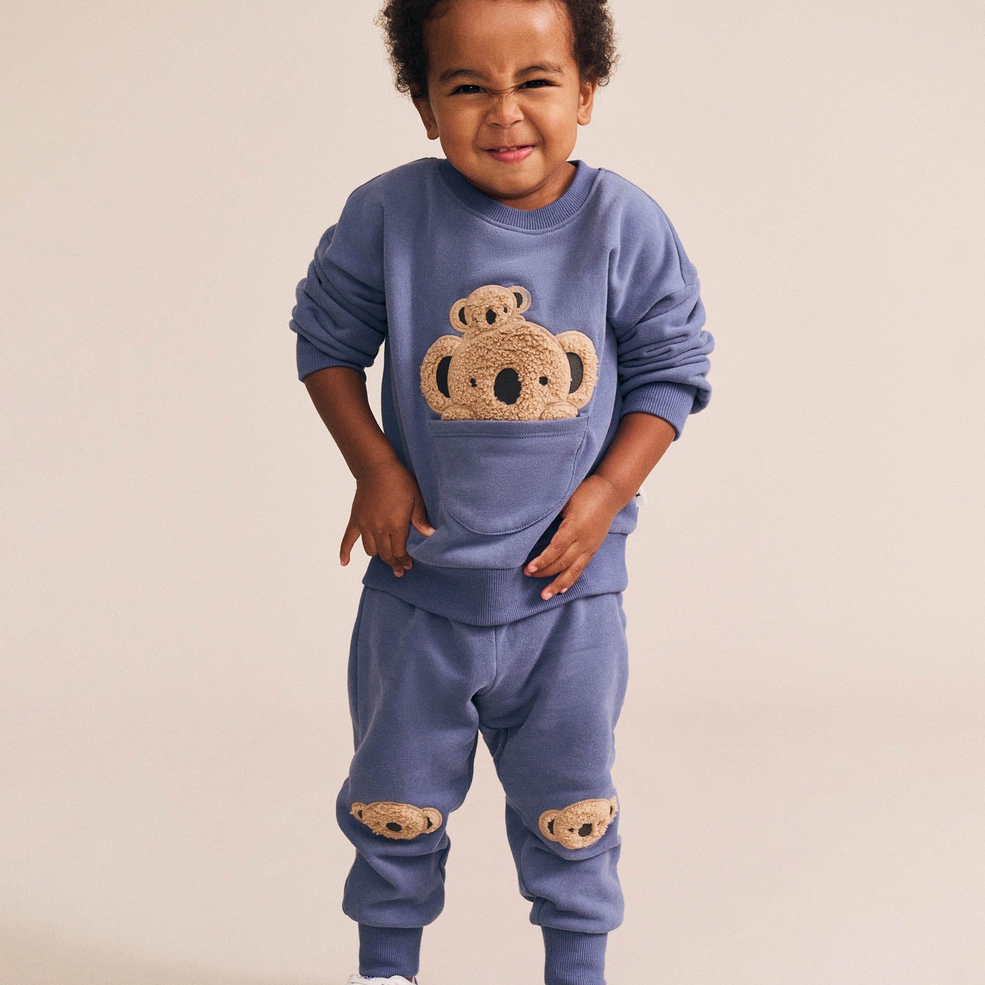 Child wearing blue Huxbaby Peekaboo Kel Pocket Sweatshirt with koala applique design in full body front view