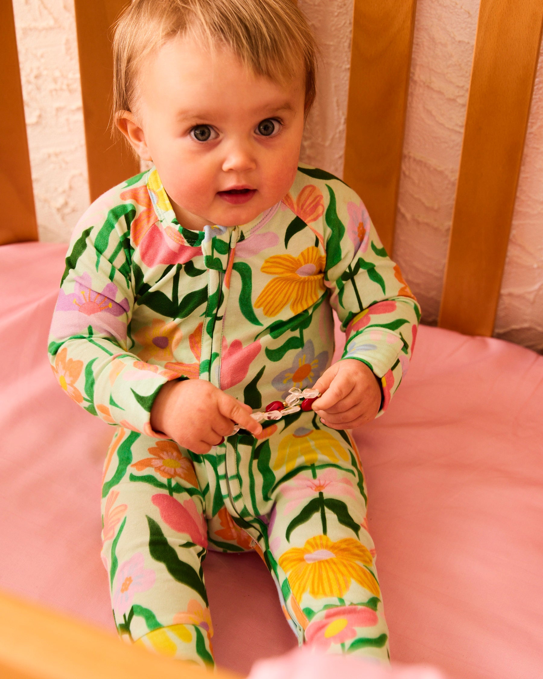 Front view of baby in Popping Flowers Green Long Sleeve Zip Romper sitting on pink bedding