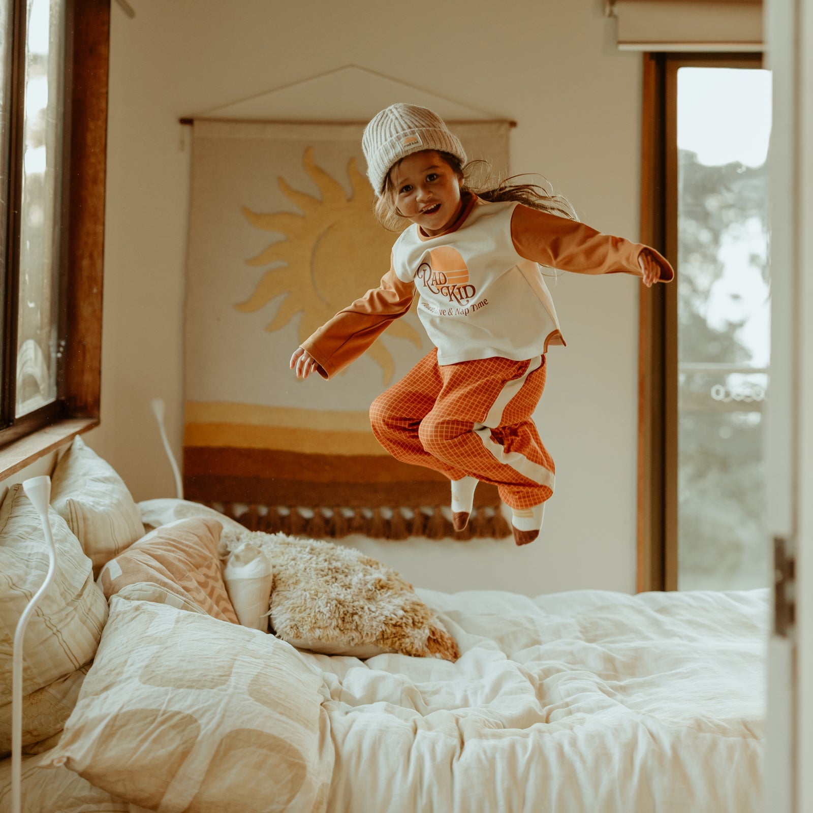 Child jumping on bed wearing Rad Kid Organic Stretch Pj Set with orange sleeves and checkered pants
