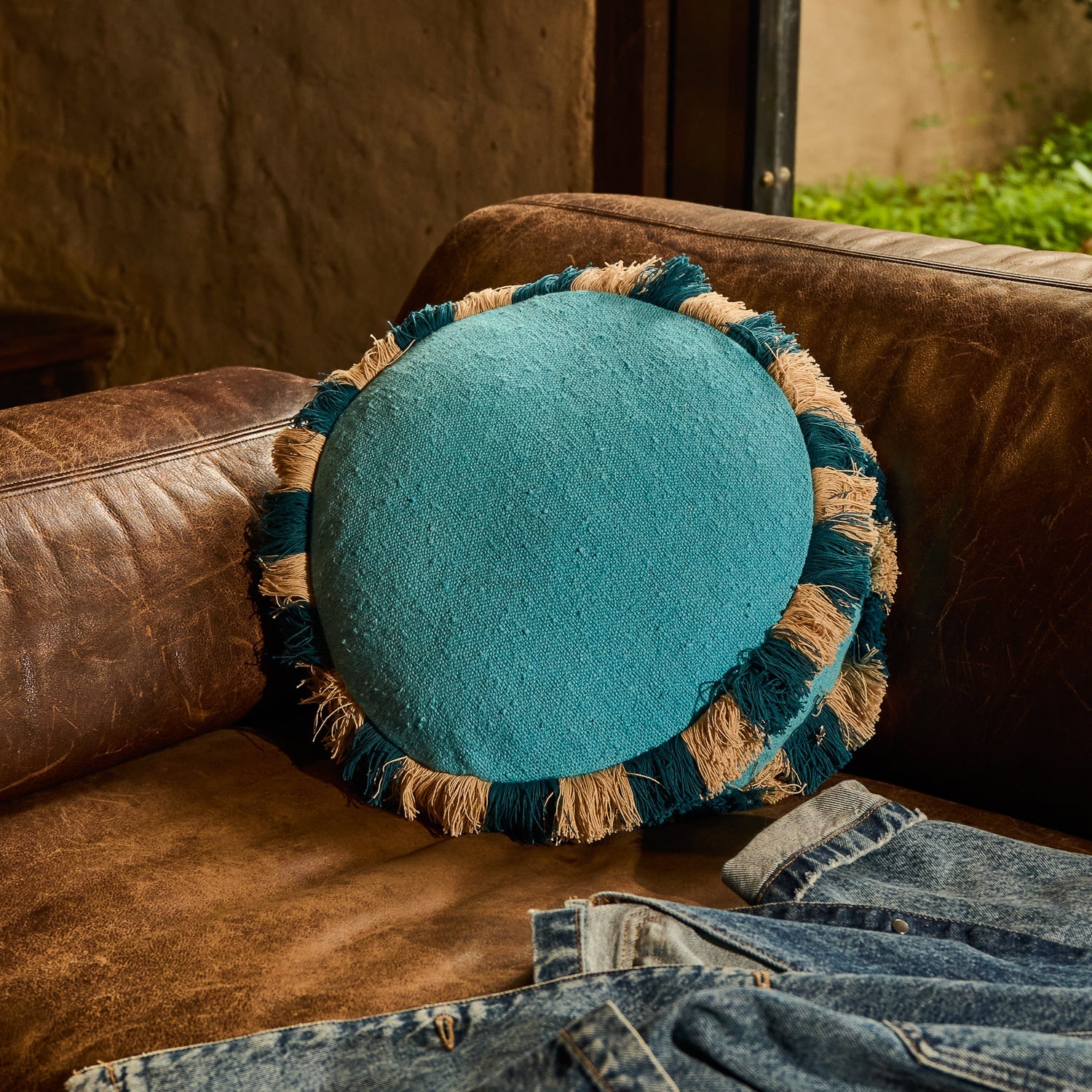 Turquoise round cushion with striped teal and natural fringe trim propped against a brown leather sofa