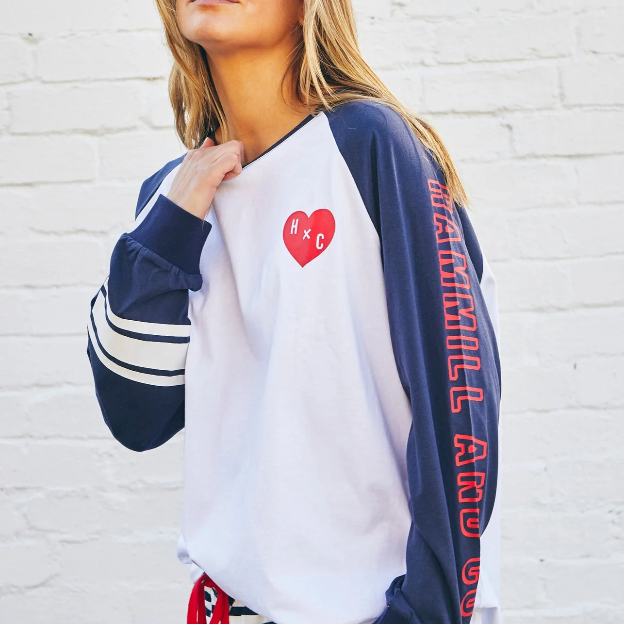 Woman wearing a navy and white long-sleeve shirt with red text and a heart design, standing against a white brick wall.