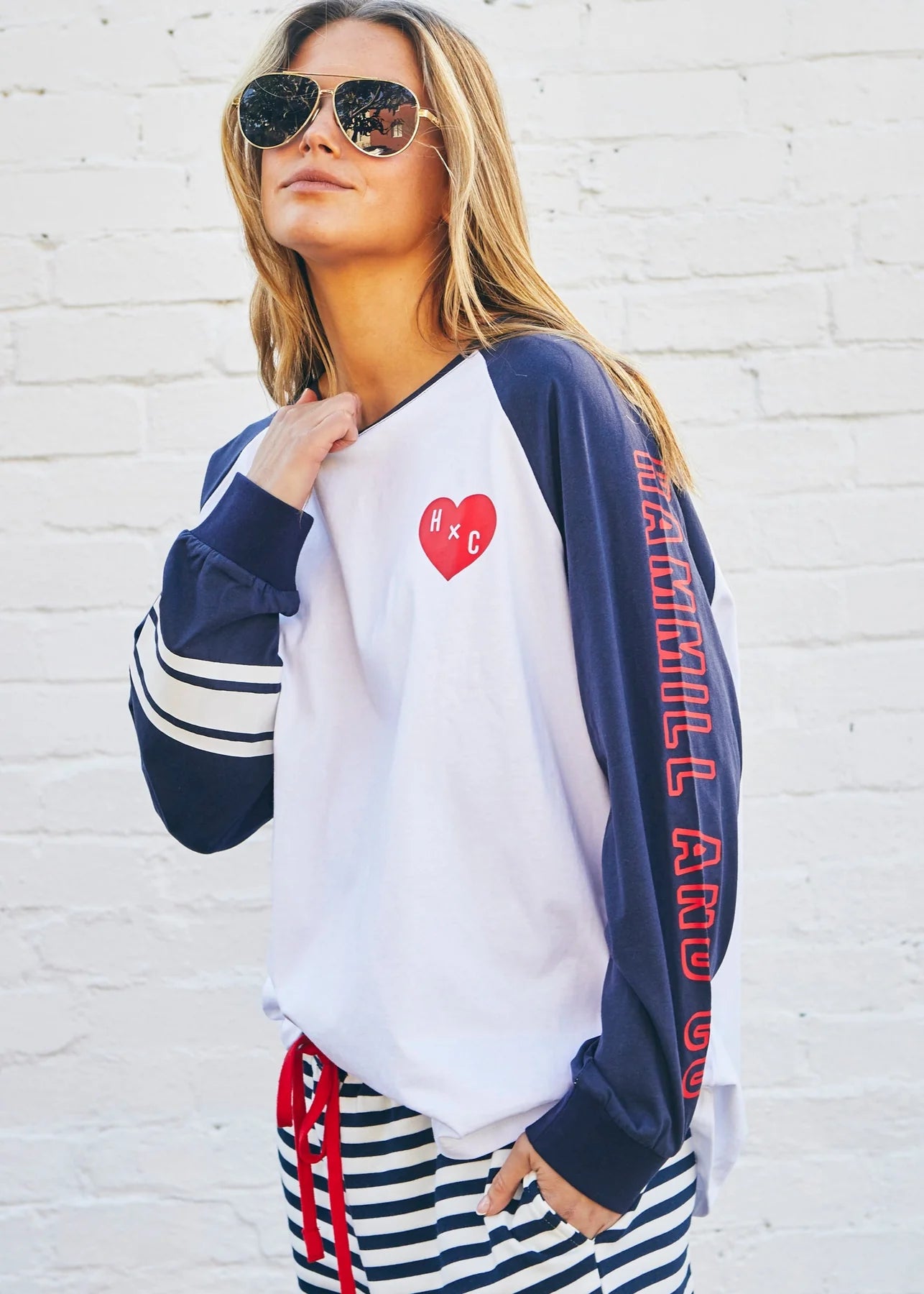 Woman wearing a navy and white long-sleeve shirt with red text and a heart design, standing against a white brick wall.