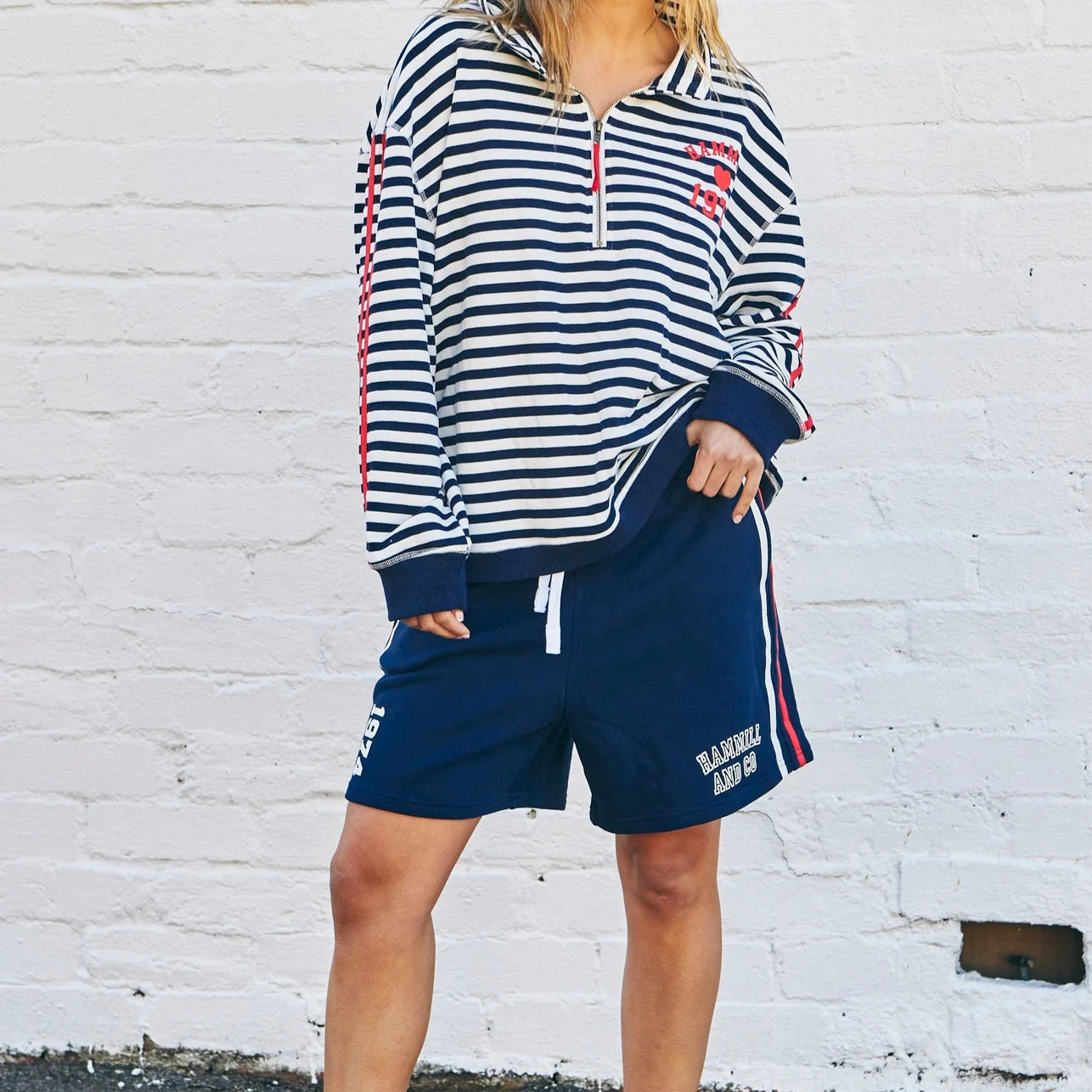 Woman wearing a striped hoodie and shorts with red accents against a white brick wall.
