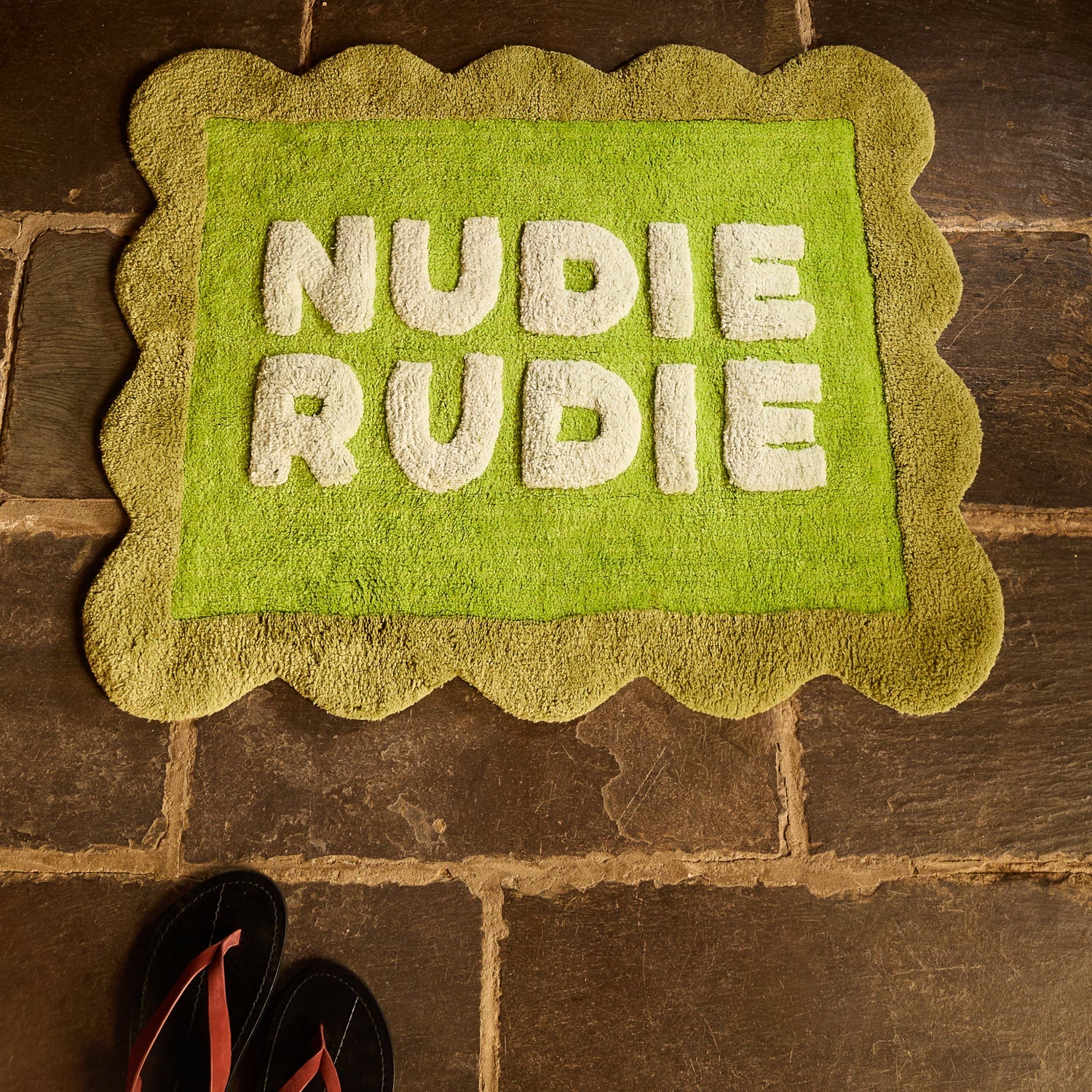 Cactus Green Nudie Rudie bath mat with scalloped border laid on stone tile floor, overhead angle