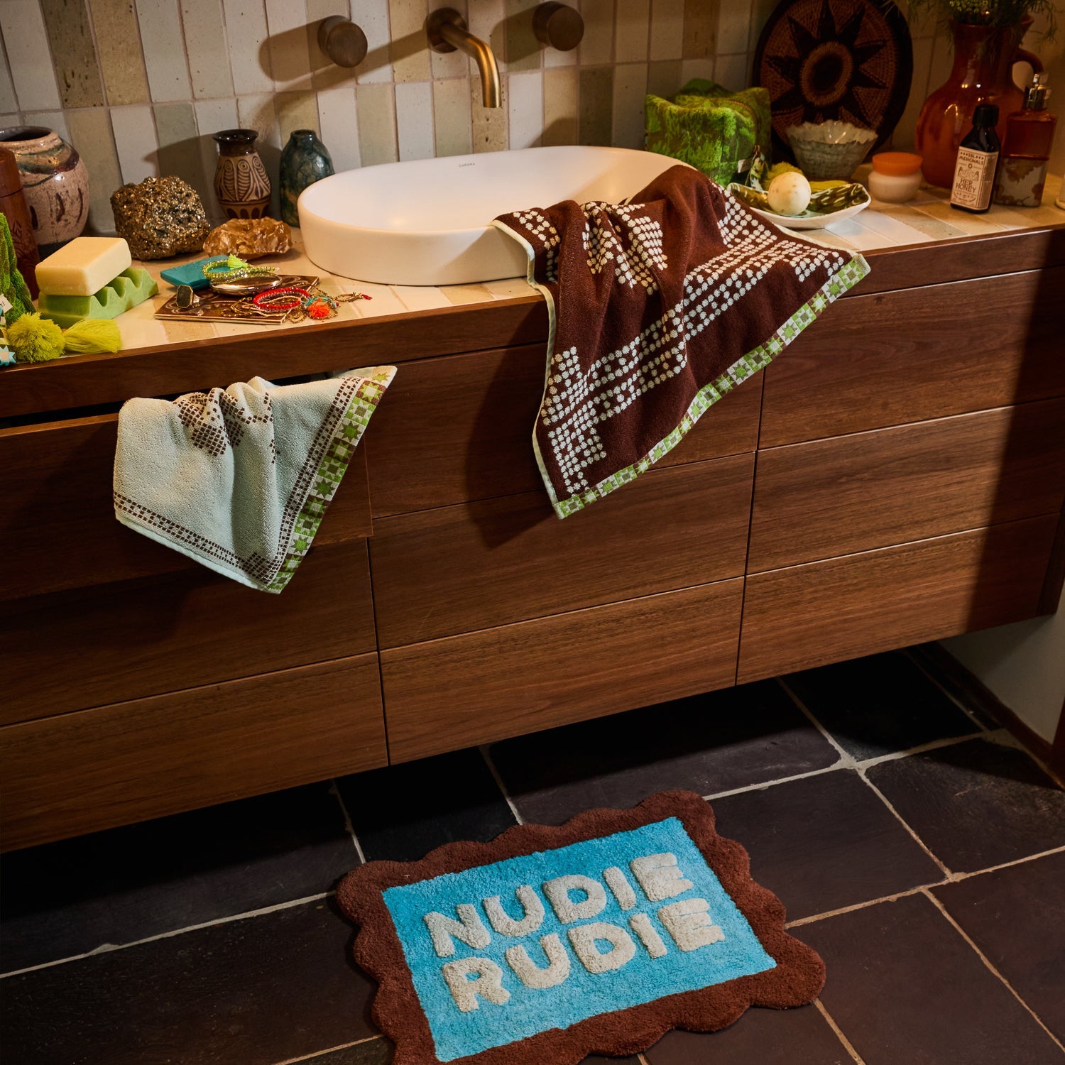 Turquoise Nudie Rudie bath mat with brown scalloped border placed in front of a wooden bathroom vanity