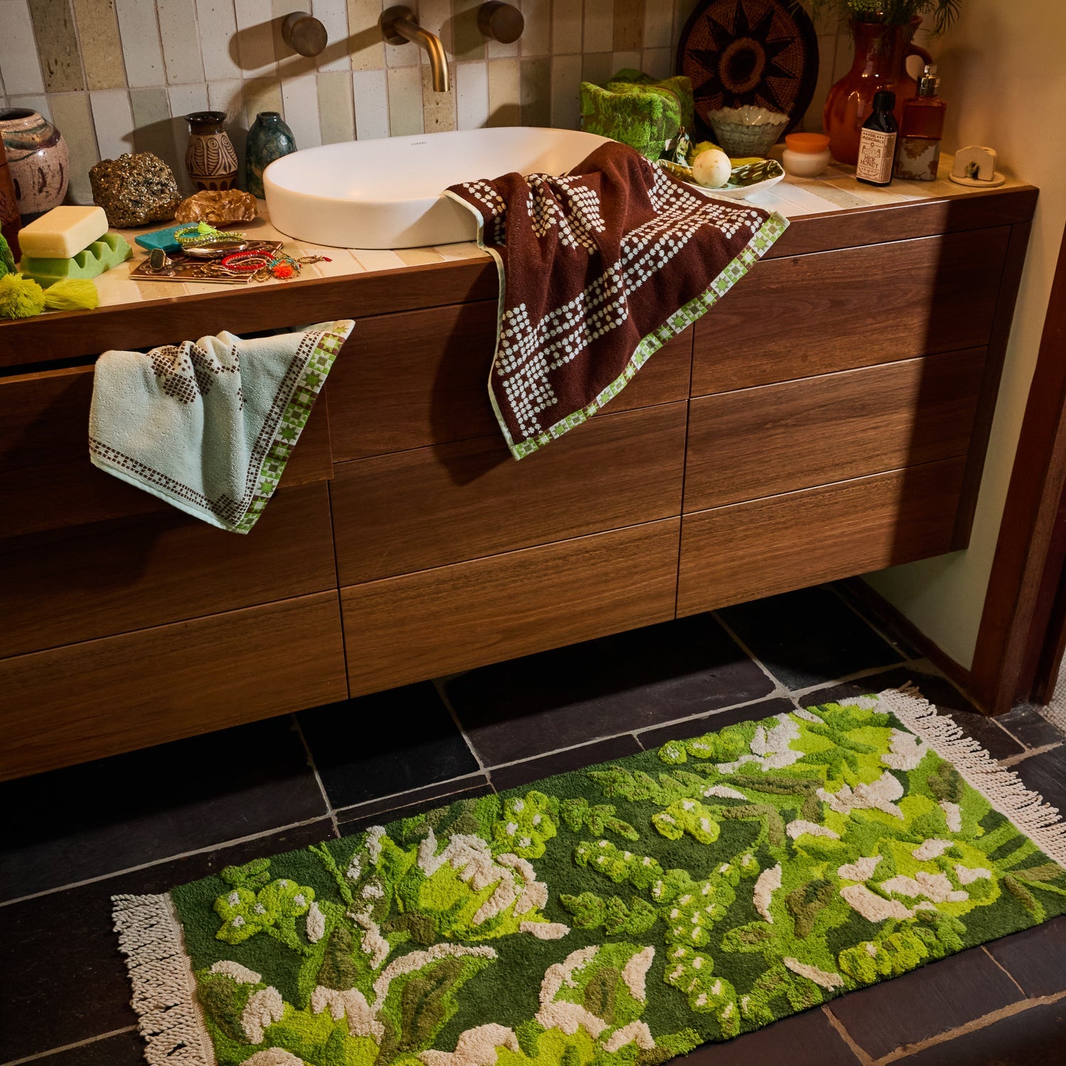 Green and white floral tufted bath runner with cream fringe styled on dark tile floor beside a wooden vanity