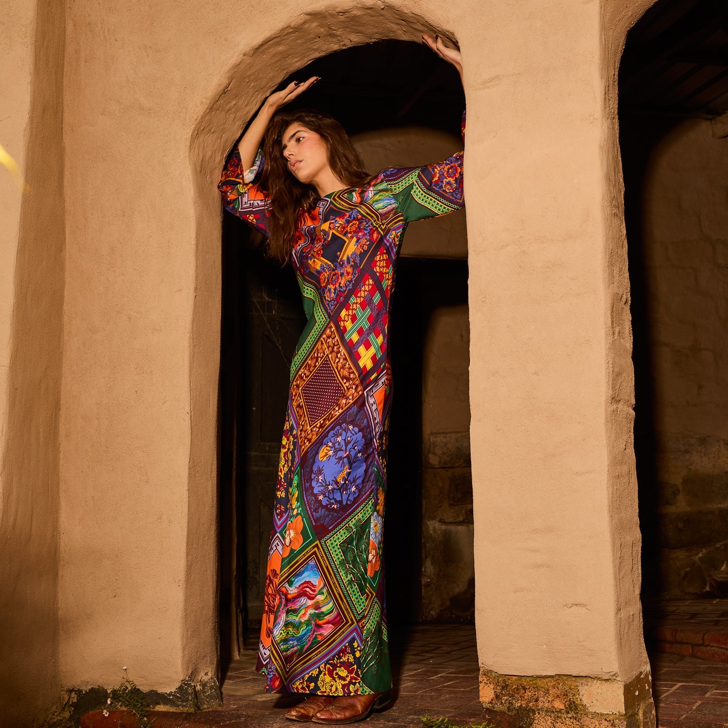 Full-length side view of a woman in a multicolour long-sleeve maxi dress standing in a stone archway
