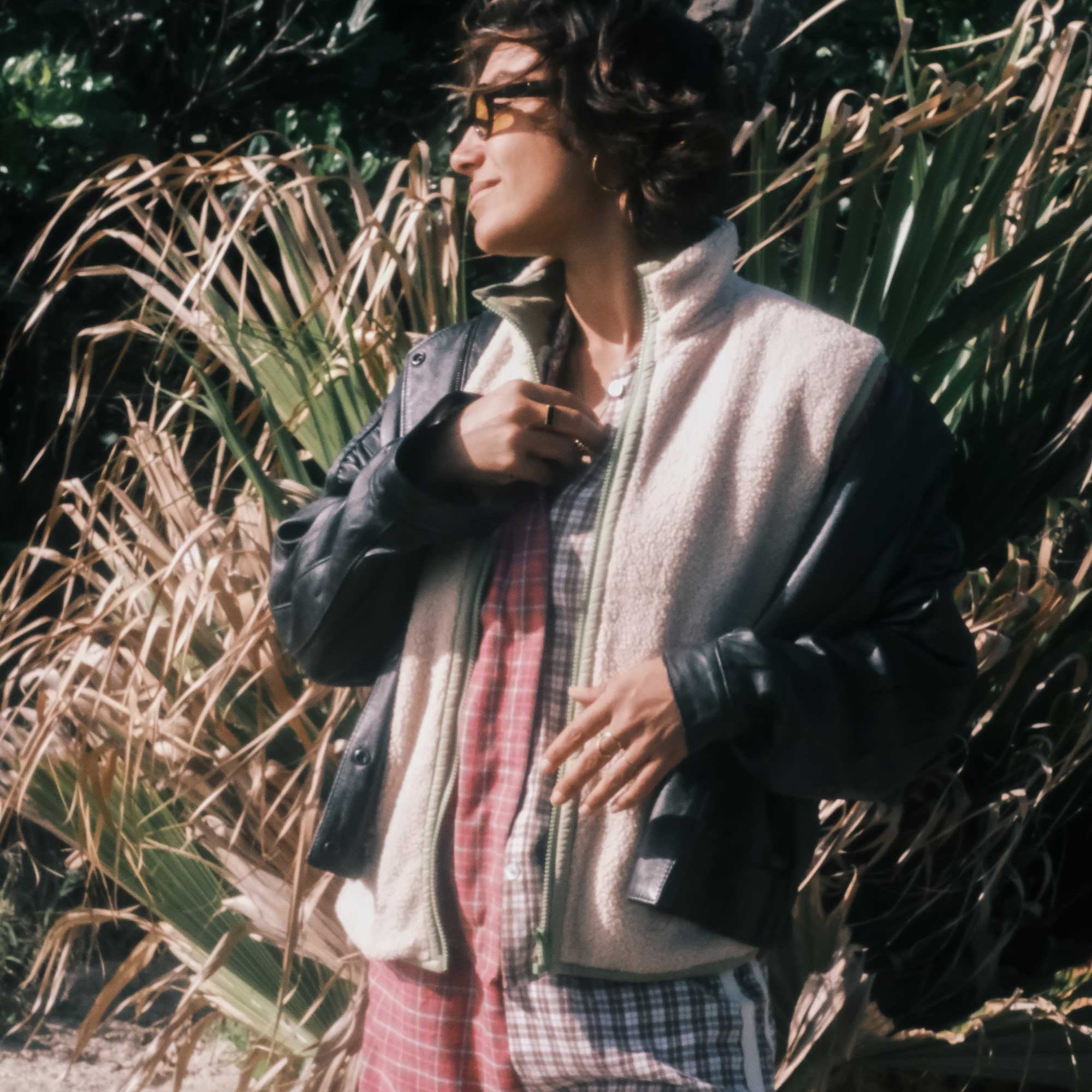 Woman wearing a cream sherpa front zip vest layered over a plaid outfit outdoors among coastal vegetation