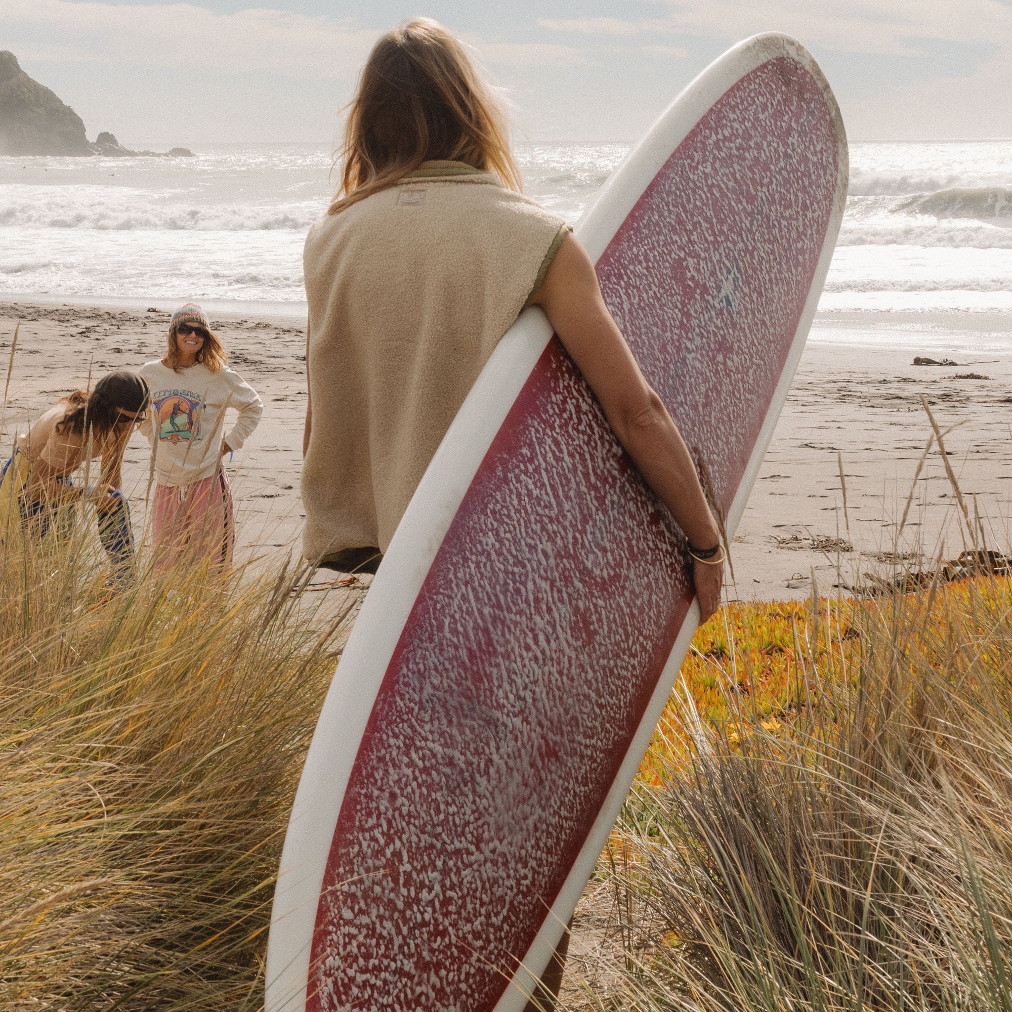 Woman in a cream sherpa front zip vest holding a surfboard on a beach, shot from behind