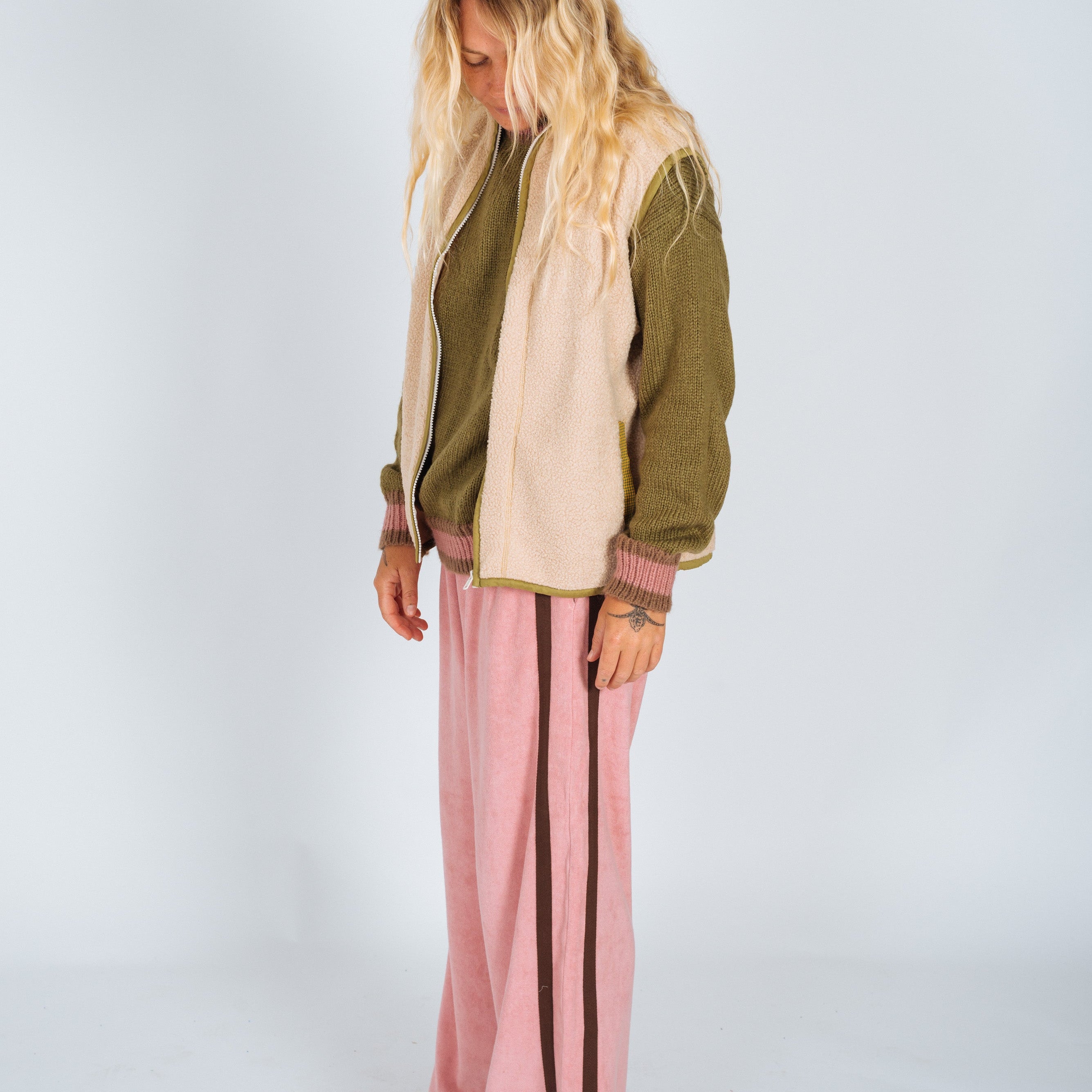 Side view of a cream sherpa front zip vest with olive trim worn over a knit sweater and pink wide-leg pants
