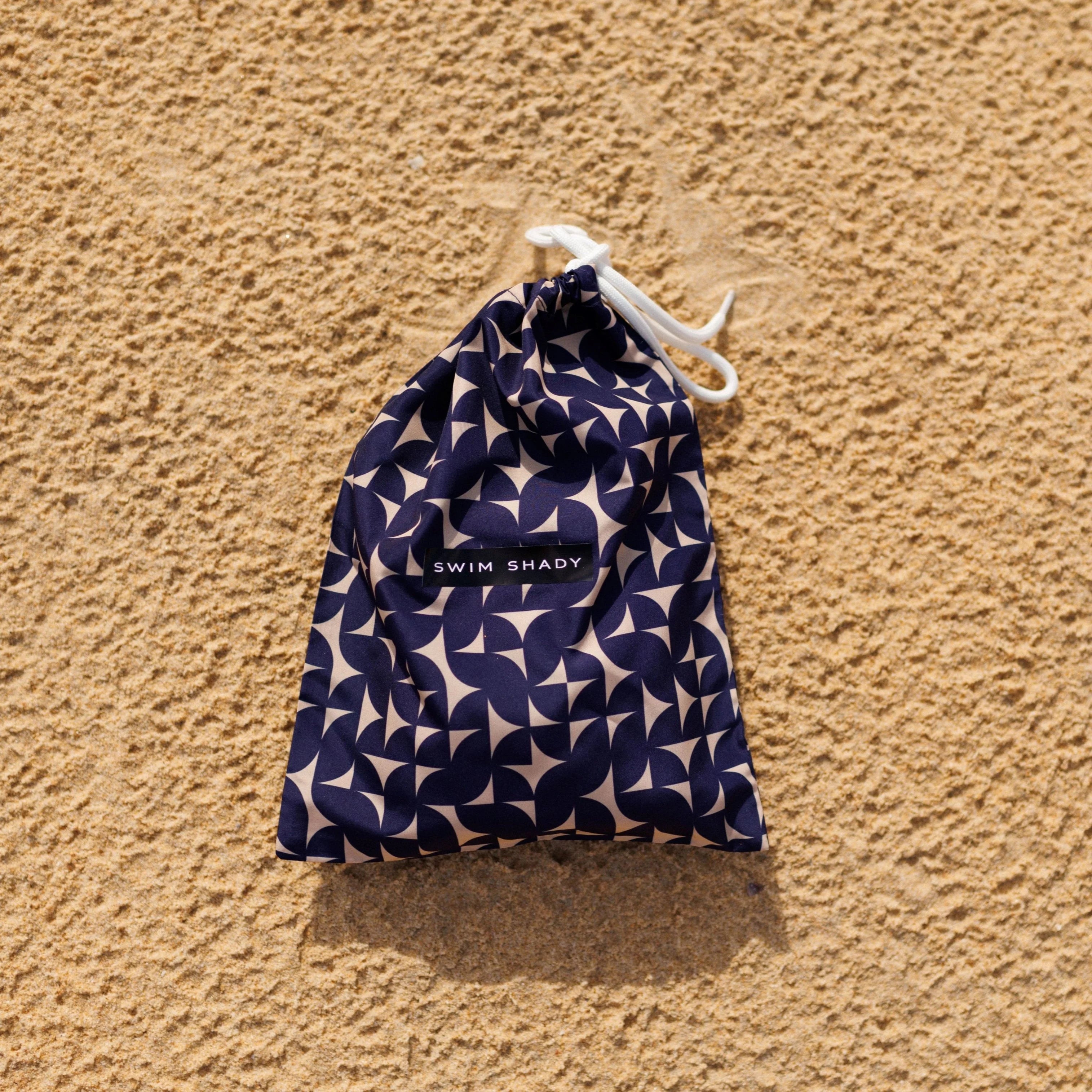 Navy patterned drawstring bag with 'Swim Shady' label on a sandy background