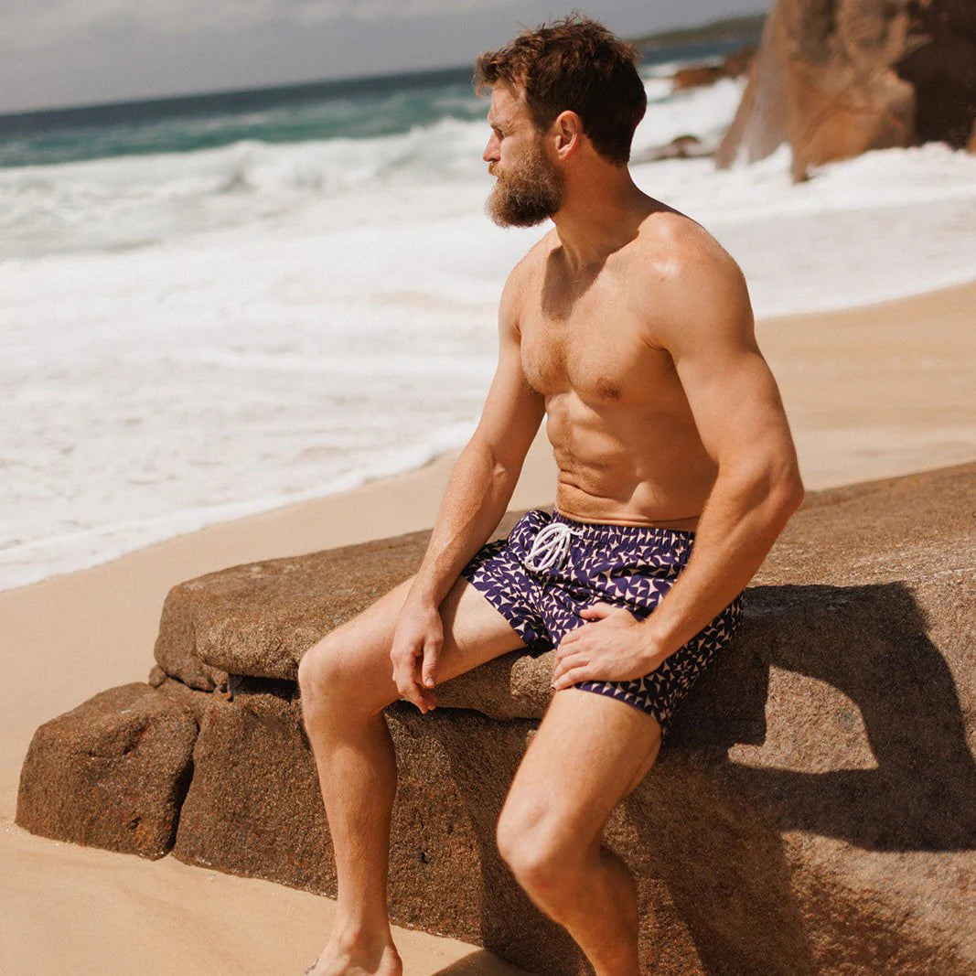 Man sitting on a rock by the ocean wearing patterned swim shorts.
