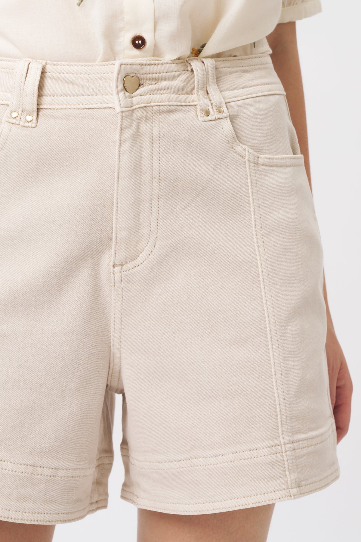 Close-up front detail of cream denim shorts showing heart-shaped gold button, front pocket, and contrast stitching