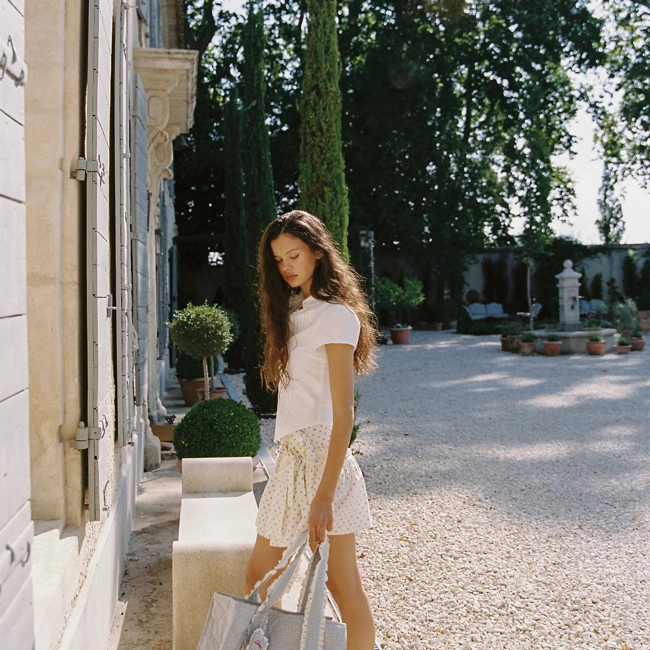 Woman holding a large bag in an outdoor setting with trees and a building.