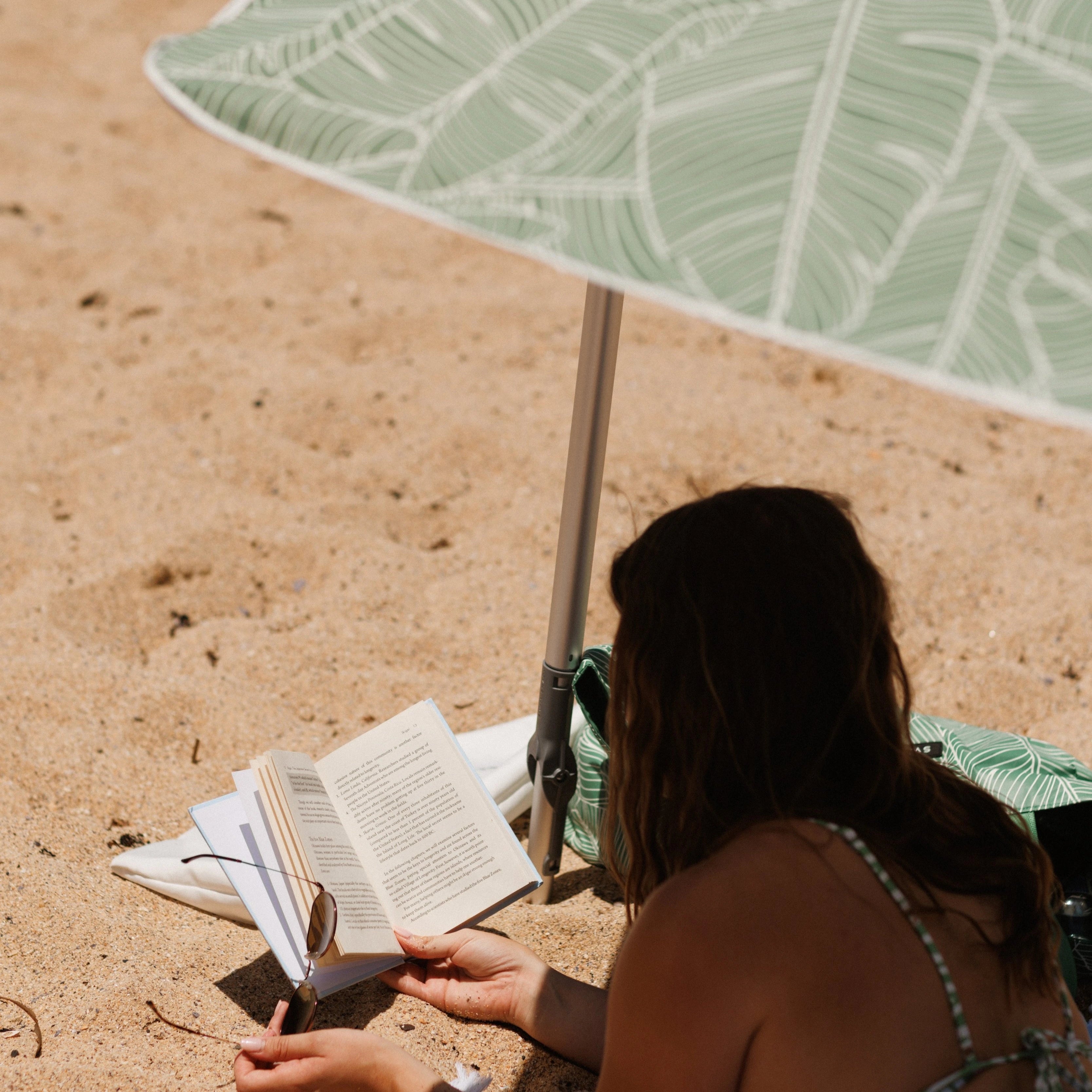 Person reading a book under a green leaf-patterned umbrella on a sandy beach.