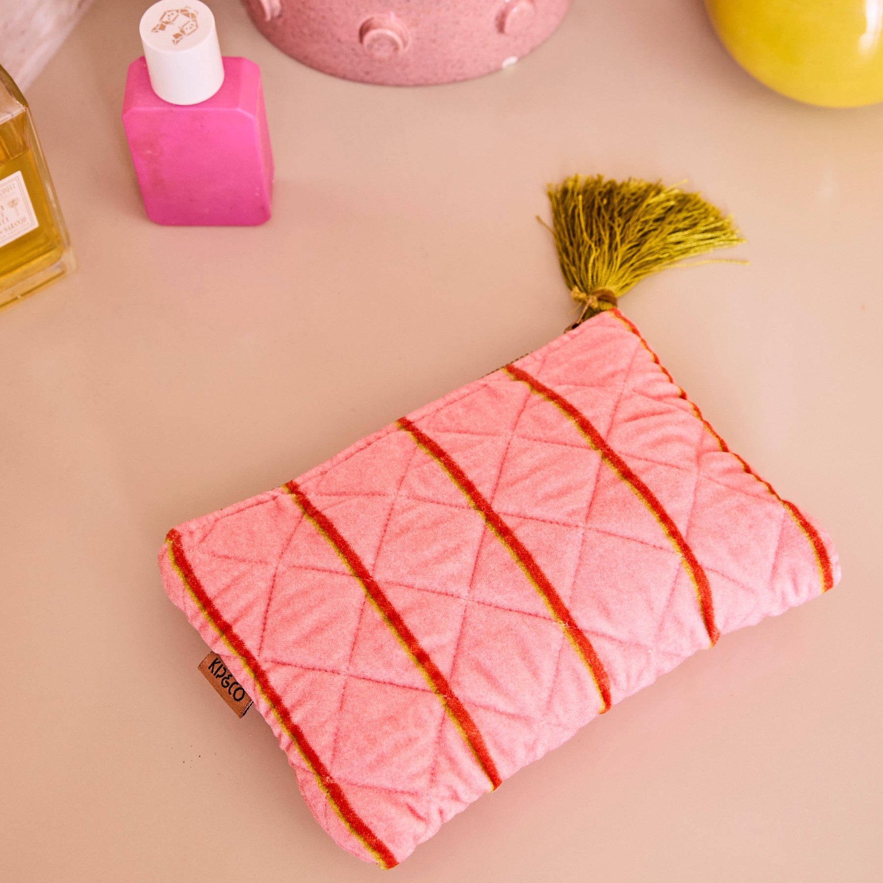 Pink velvet Wategos Soiree Stripe cosmetics purse by Kip & Co with red stripes and green tassel on beige surface