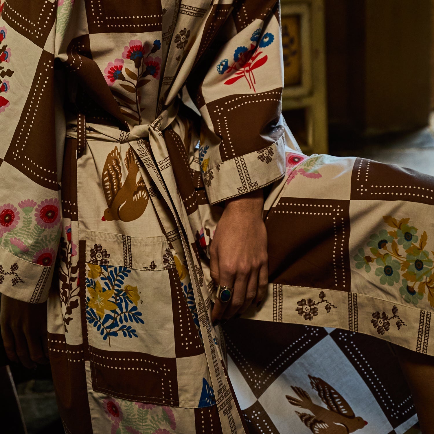 Close-up of cotton robe showing brown and cream checkered print with bird and floral motifs, tie belt and patch pocket detail