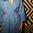 Close-up of light blue denim robe showing red cross-stitch flower embroidery, V-neckline, and belted waist detail