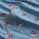 Close-up flat detail of light blue denim robe pocket area with red cross-stitch poppy flower embroidery